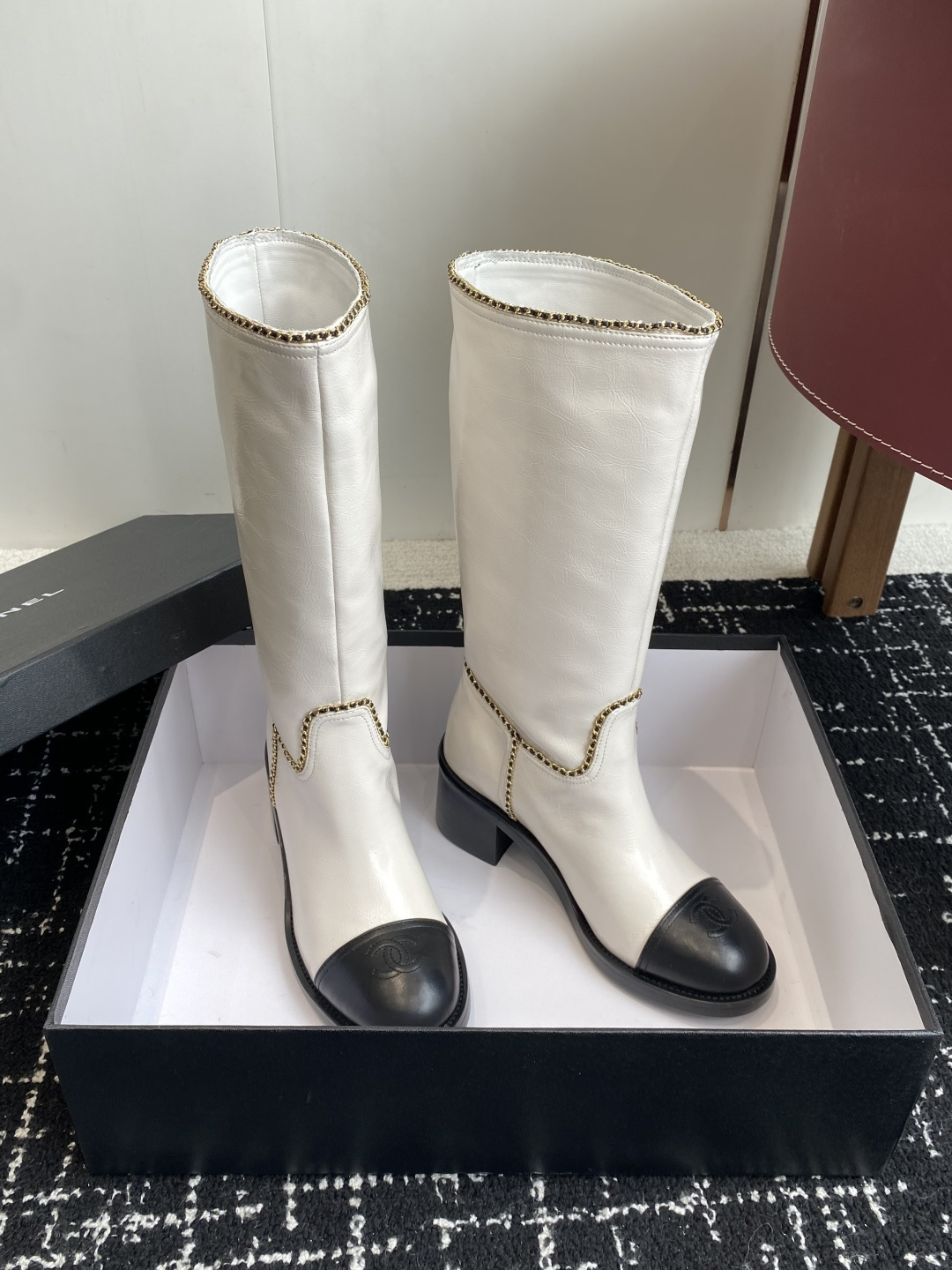 Pair of luxury white and black two-tone knee-high boots with gold chain trim and block heels on a black display box. - Hình 3