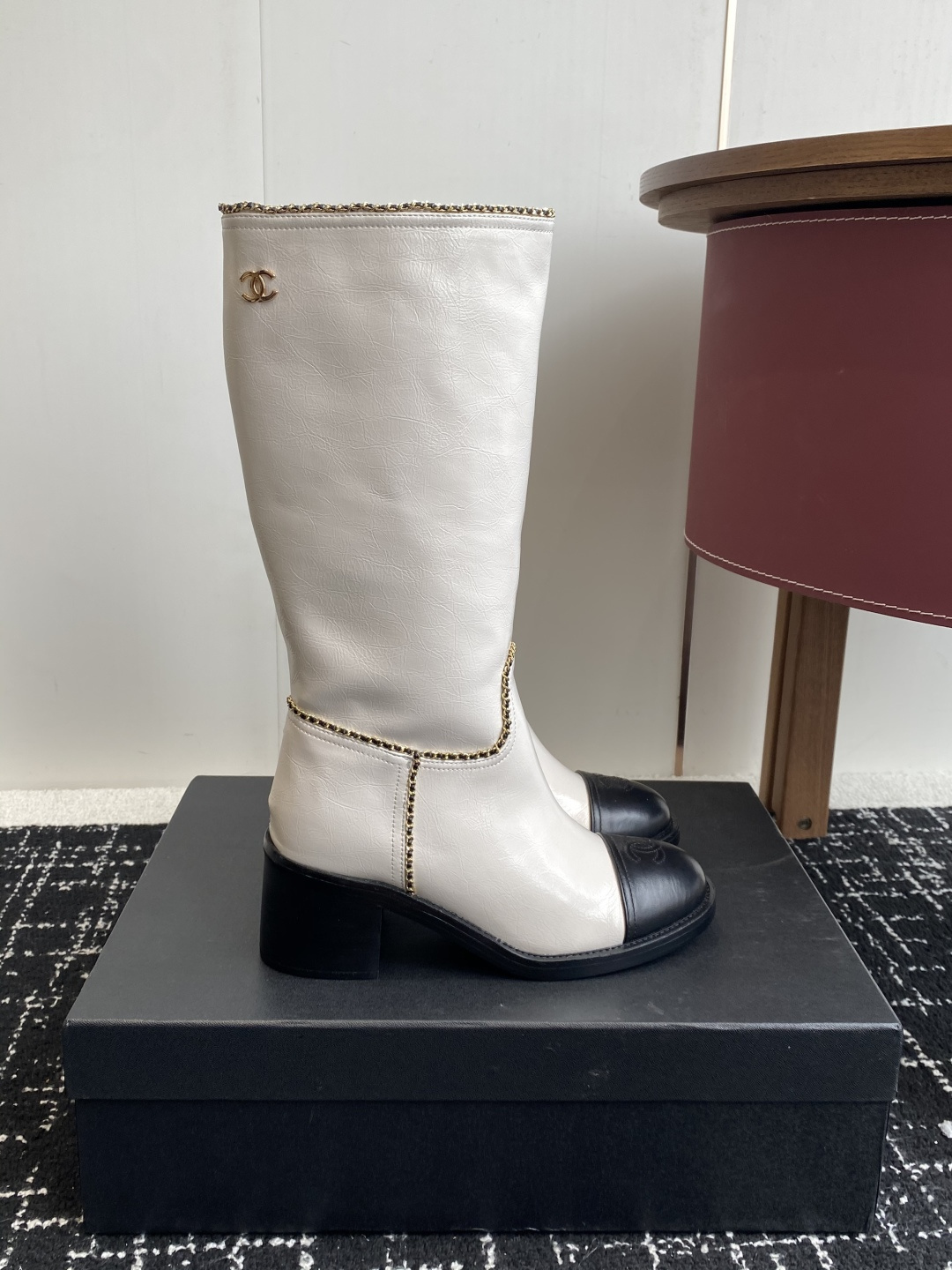 Pair of luxury white and black two-tone knee-high boots with gold chain trim and block heels on a black display box. - Hình 2