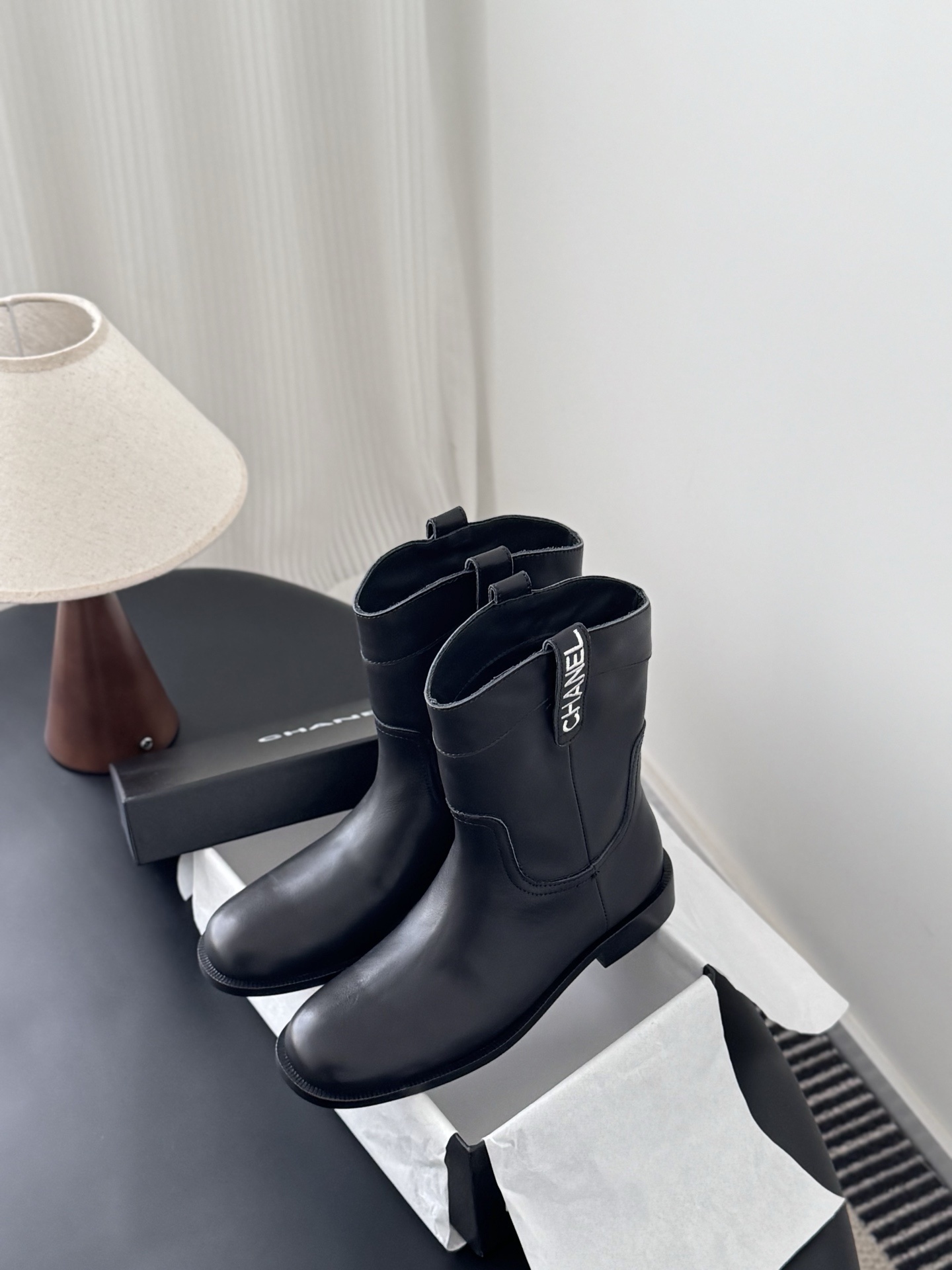 A pair of Chanel black leather mid-calf boots with silver logo hardware on the pull tabs, displayed next to a Chanel designer box. - Hình 9