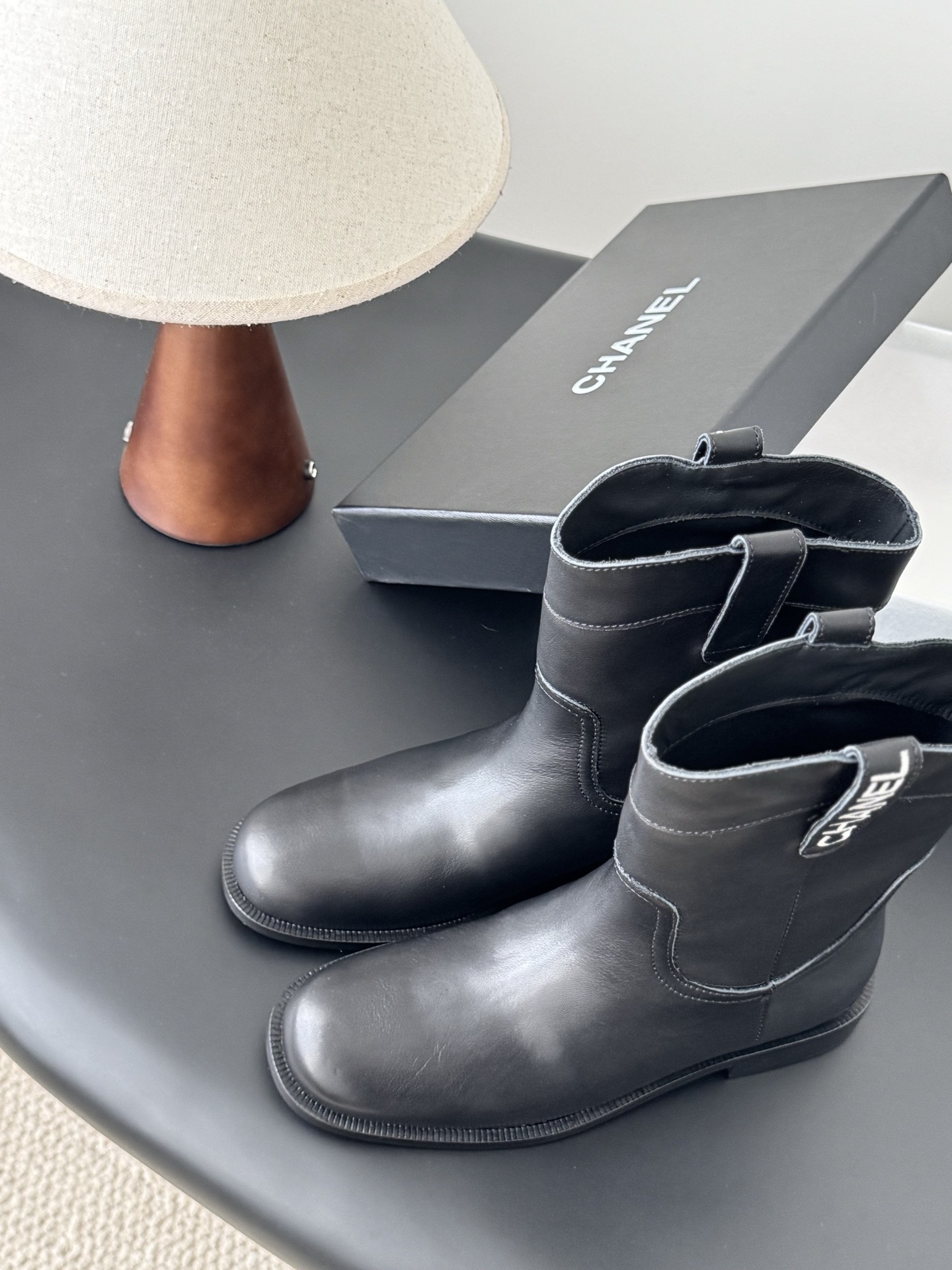 A pair of Chanel black leather mid-calf boots with silver logo hardware on the pull tabs, displayed next to a Chanel designer box. - Hình 8