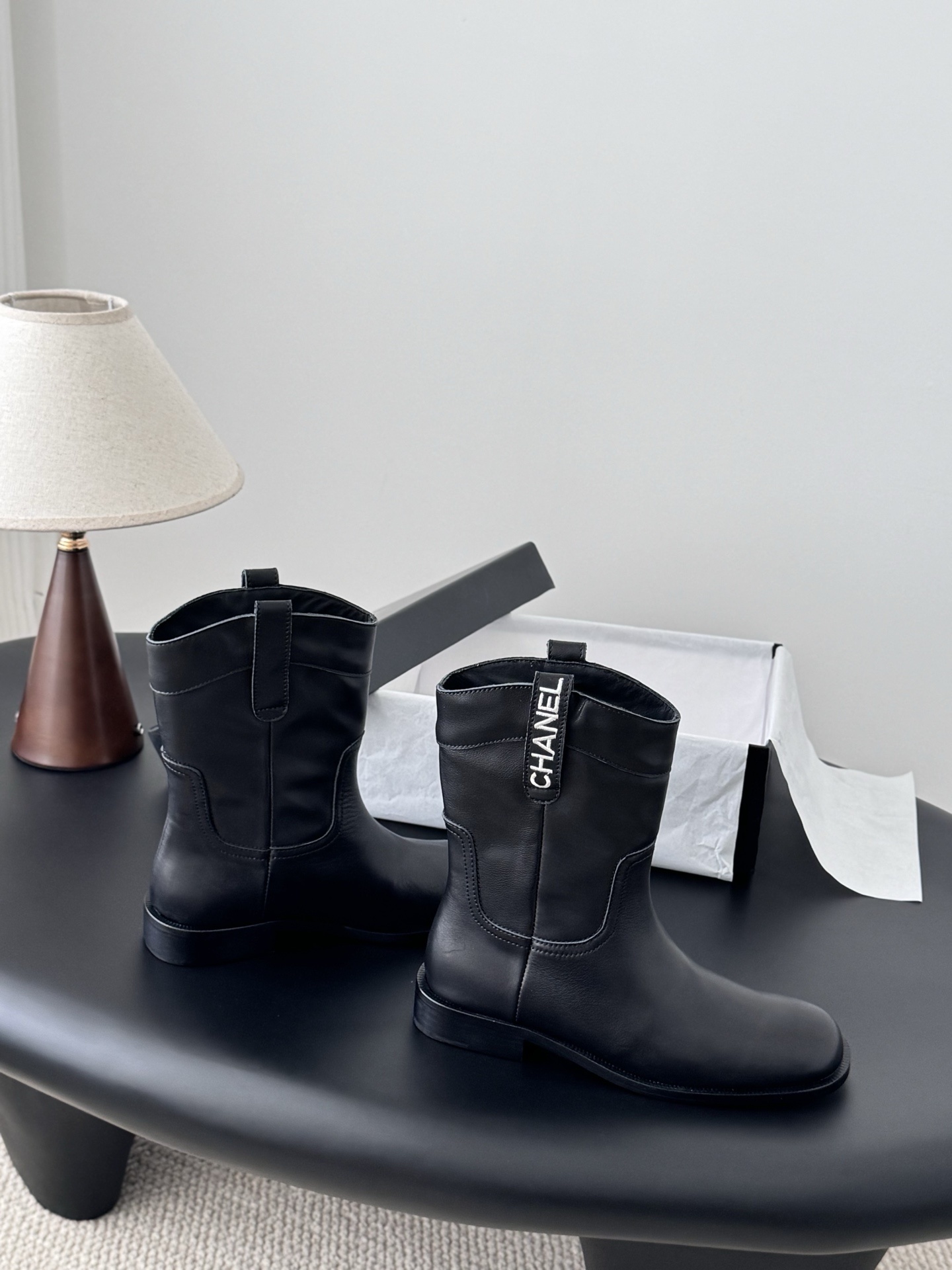 A pair of Chanel black leather mid-calf boots with silver logo hardware on the pull tabs, displayed next to a Chanel designer box. - Hình 7