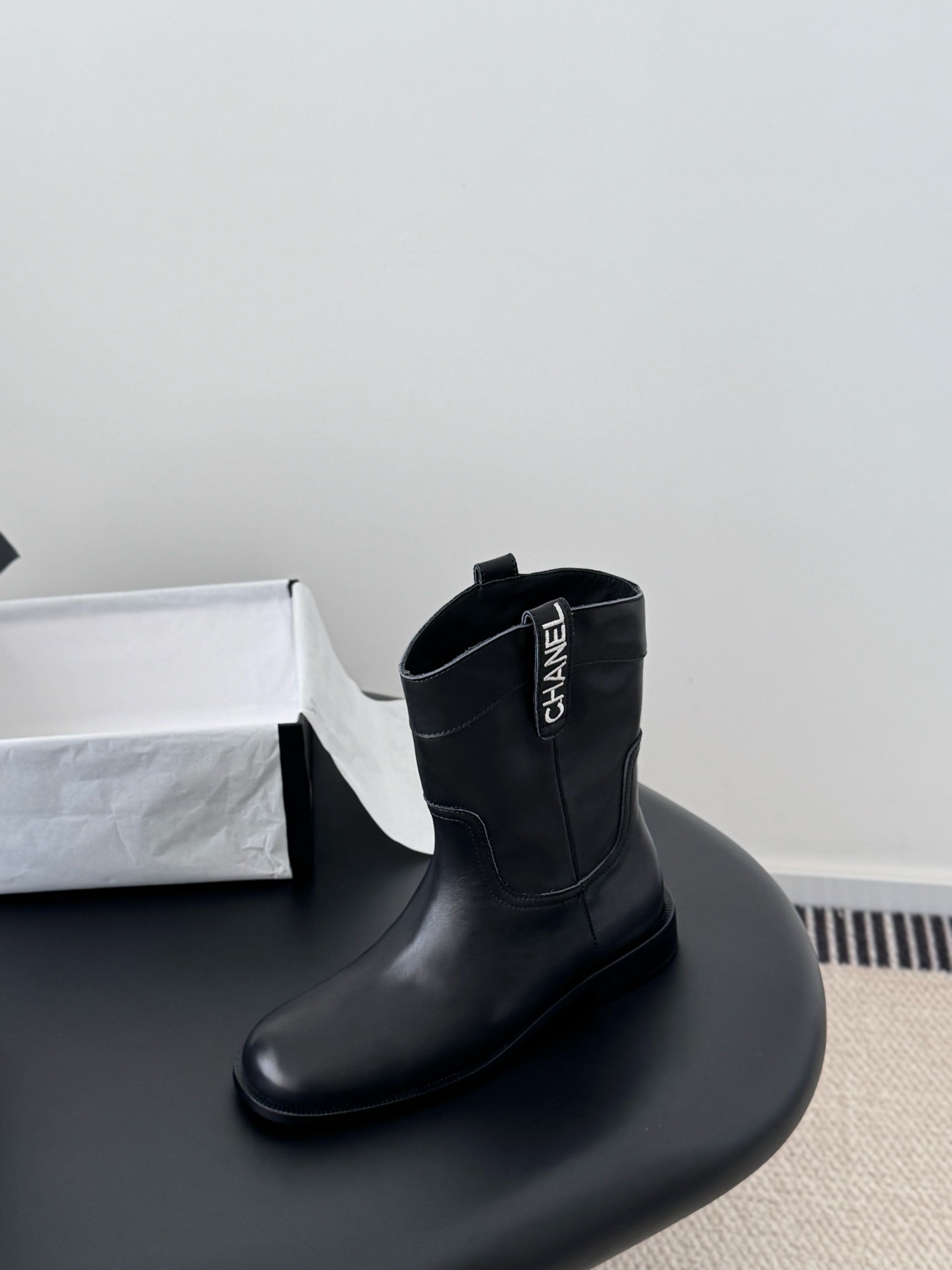 A pair of Chanel black leather mid-calf boots with silver logo hardware on the pull tabs, displayed next to a Chanel designer box. - Hình 5