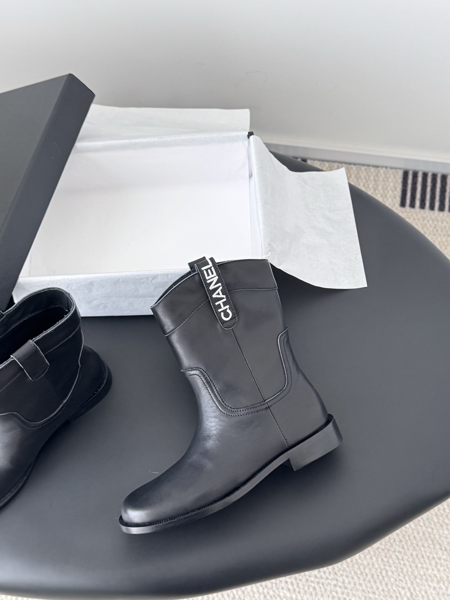 A pair of Chanel black leather mid-calf boots with silver logo hardware on the pull tabs, displayed next to a Chanel designer box. - Hình 3