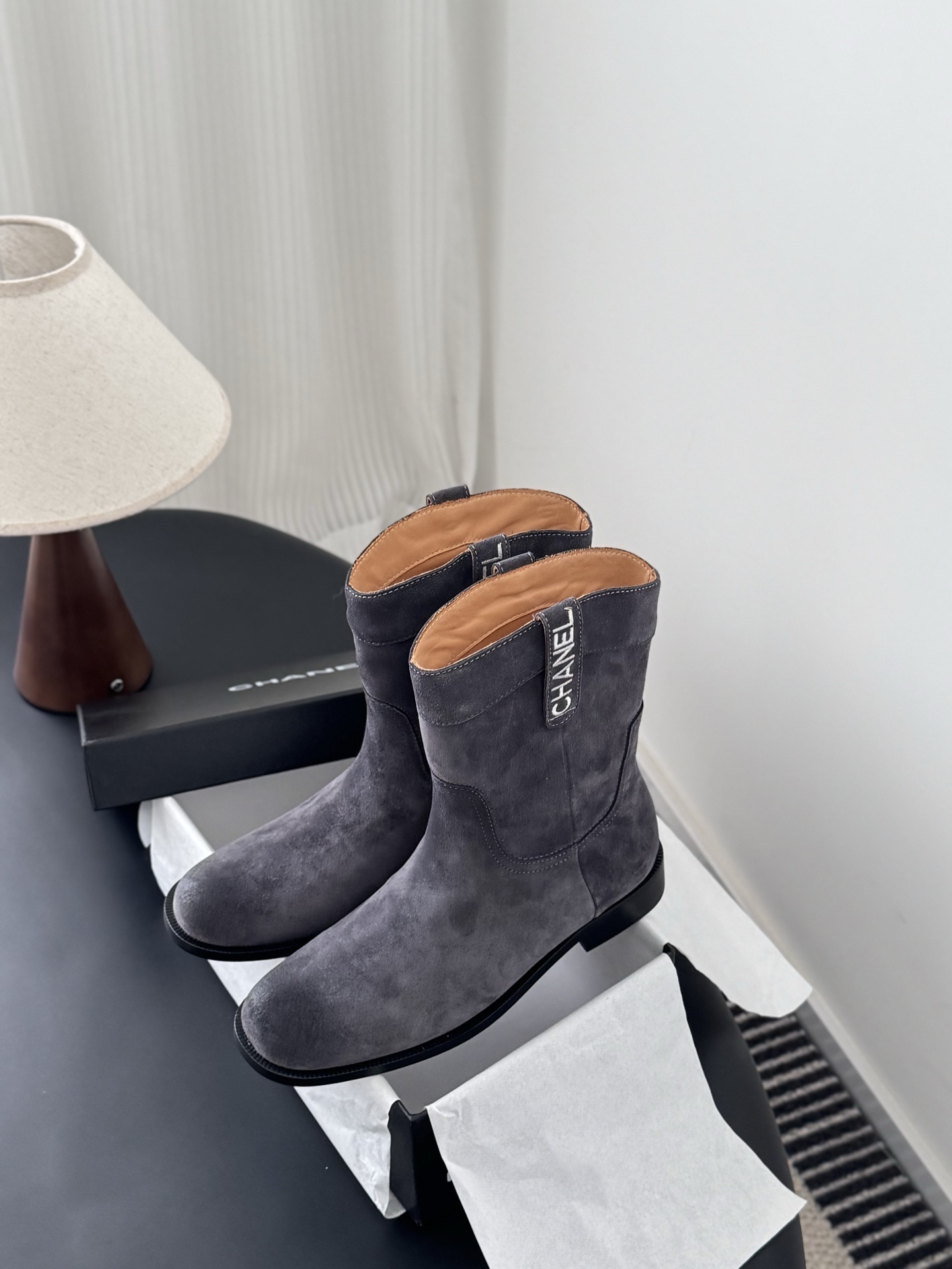 A pair of luxury Chanel grey suede mid-calf boots with logo pull tabs displayed on a black table next to their original box. - Hình 9