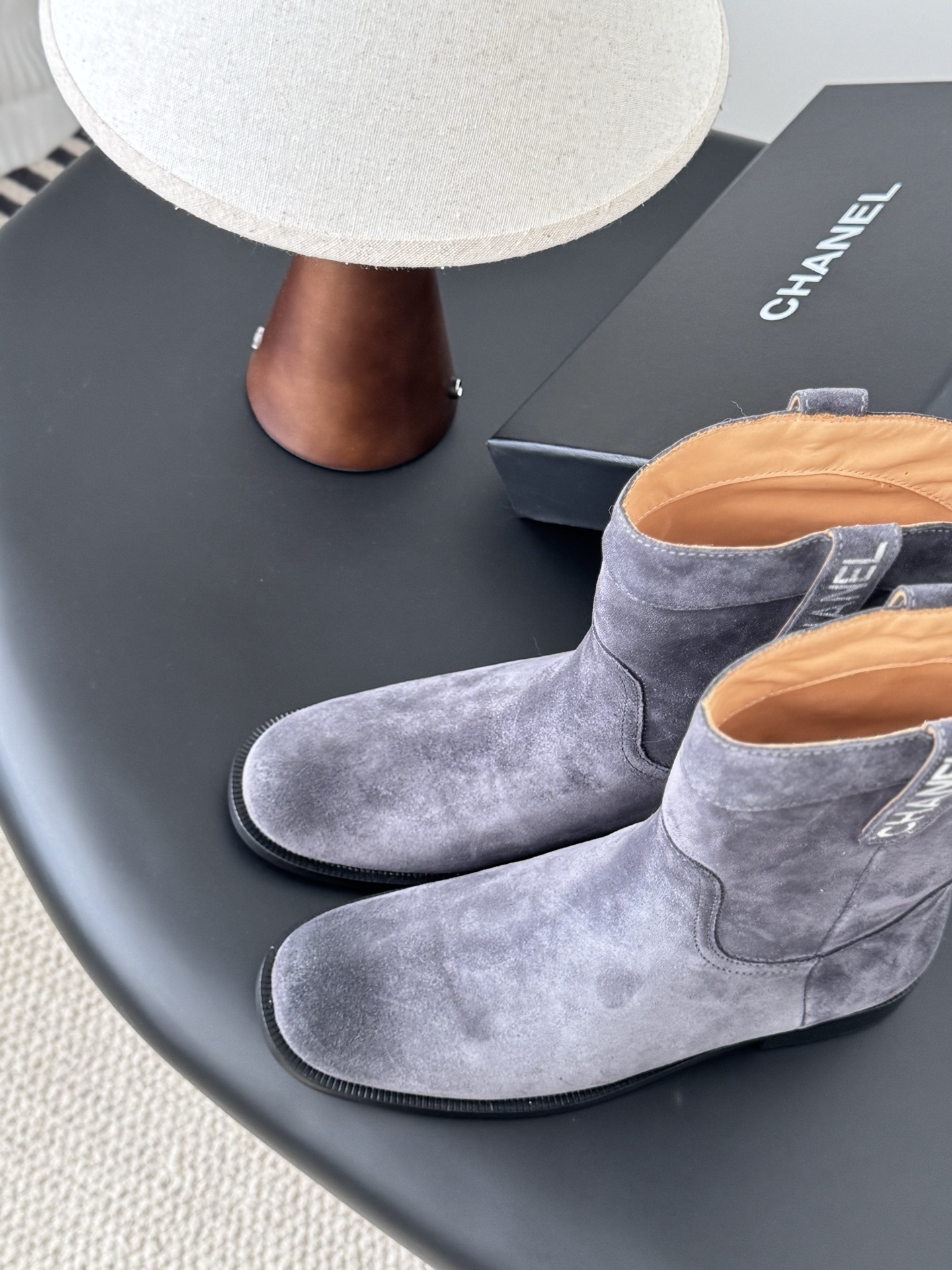 A pair of luxury Chanel grey suede mid-calf boots with logo pull tabs displayed on a black table next to their original box. - Hình 8