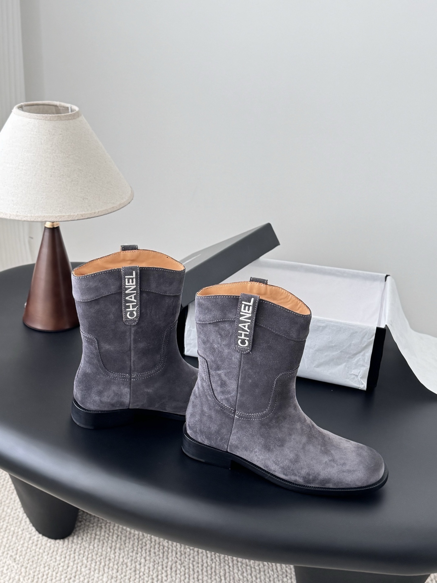 A pair of luxury Chanel grey suede mid-calf boots with logo pull tabs displayed on a black table next to their original box. - Hình 7