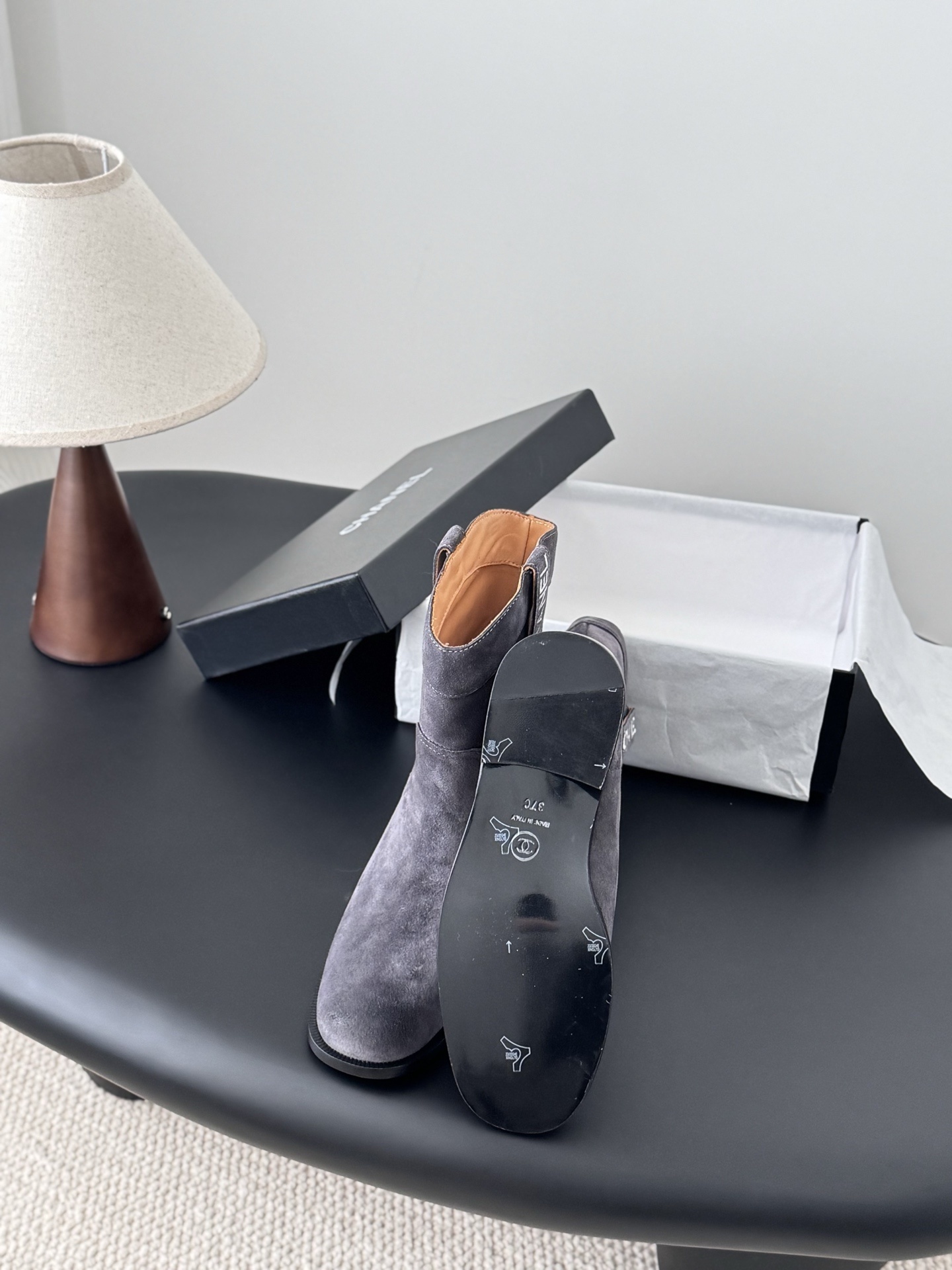 A pair of luxury Chanel grey suede mid-calf boots with logo pull tabs displayed on a black table next to their original box. - Hình 4