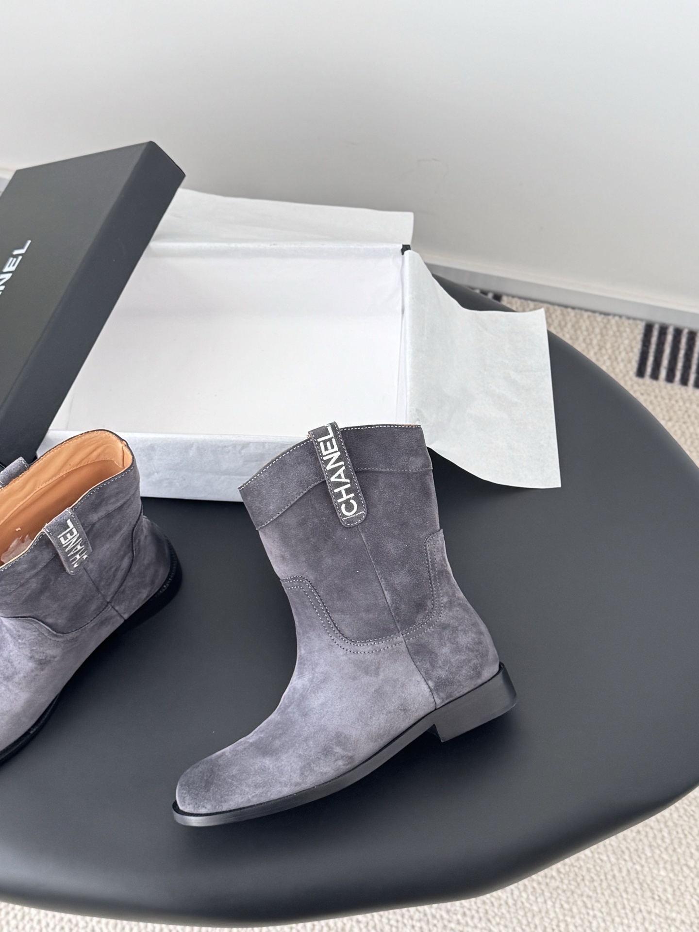 A pair of luxury Chanel grey suede mid-calf boots with logo pull tabs displayed on a black table next to their original box. - Hình 3