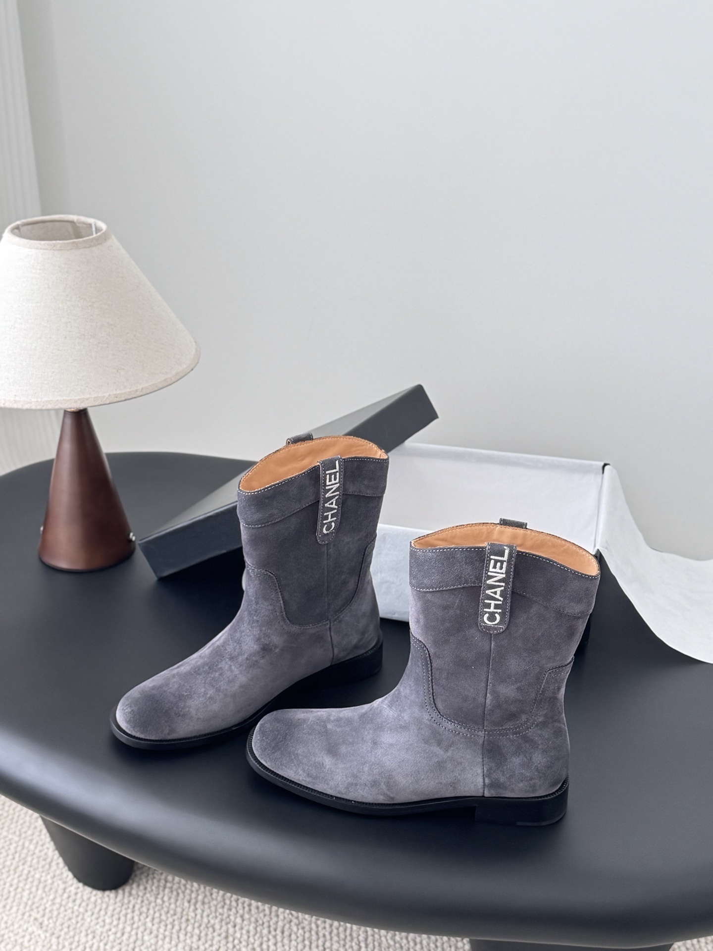 A pair of luxury Chanel grey suede mid-calf boots with logo pull tabs displayed on a black table next to their original box. - Hình 2