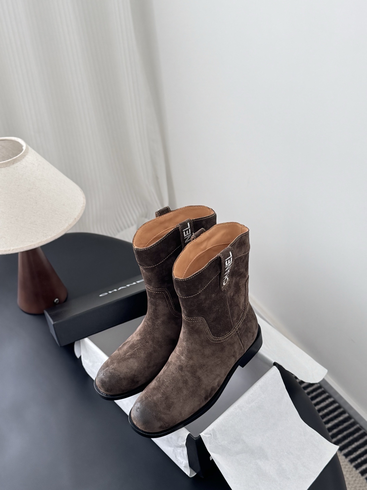A pair of Chanel brown suede western-style ankle boots with iconic pull tabs and distressed toes on a black table. - Hình 9