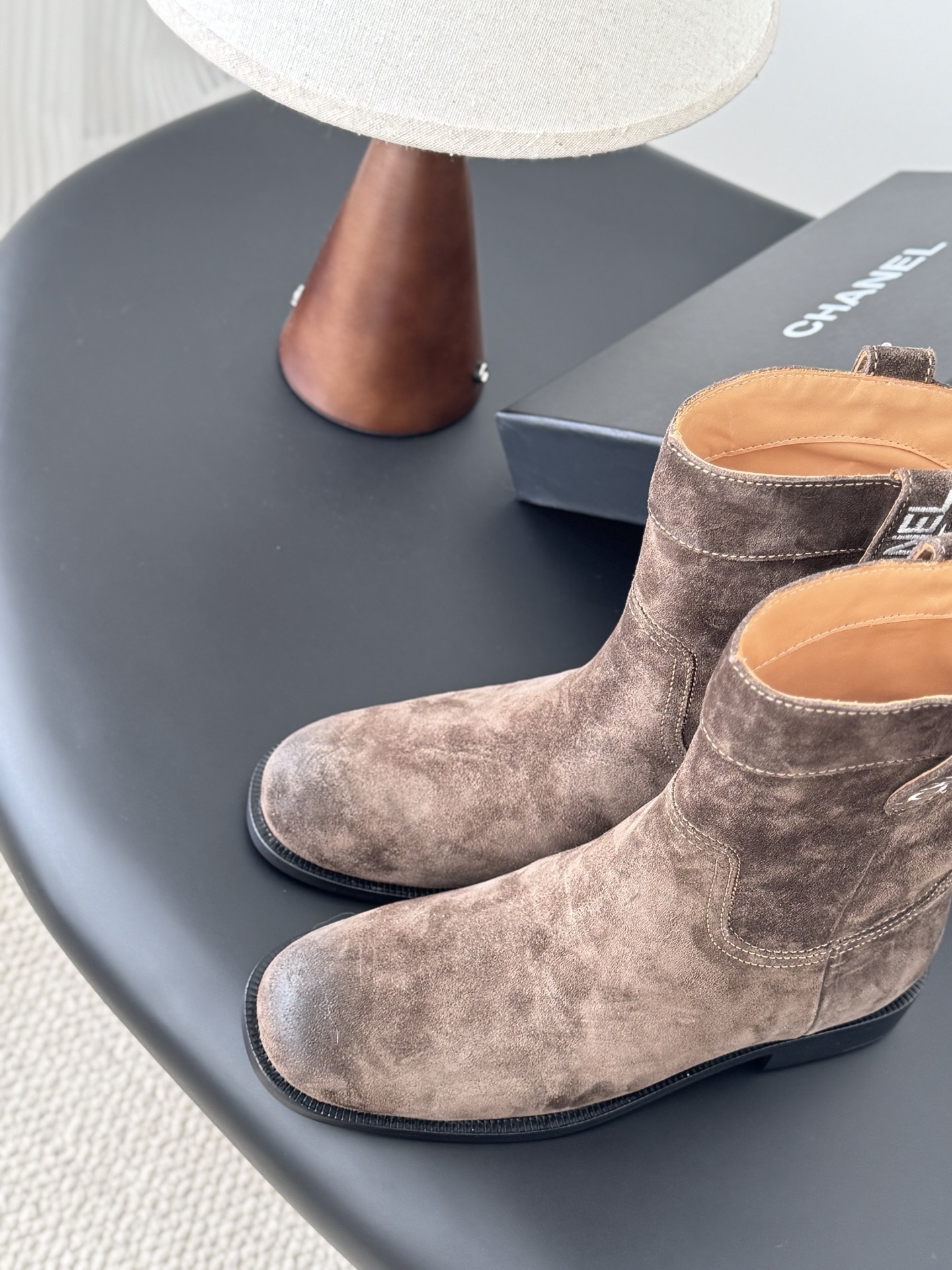 A pair of Chanel brown suede western-style ankle boots with iconic pull tabs and distressed toes on a black table. - Hình 7