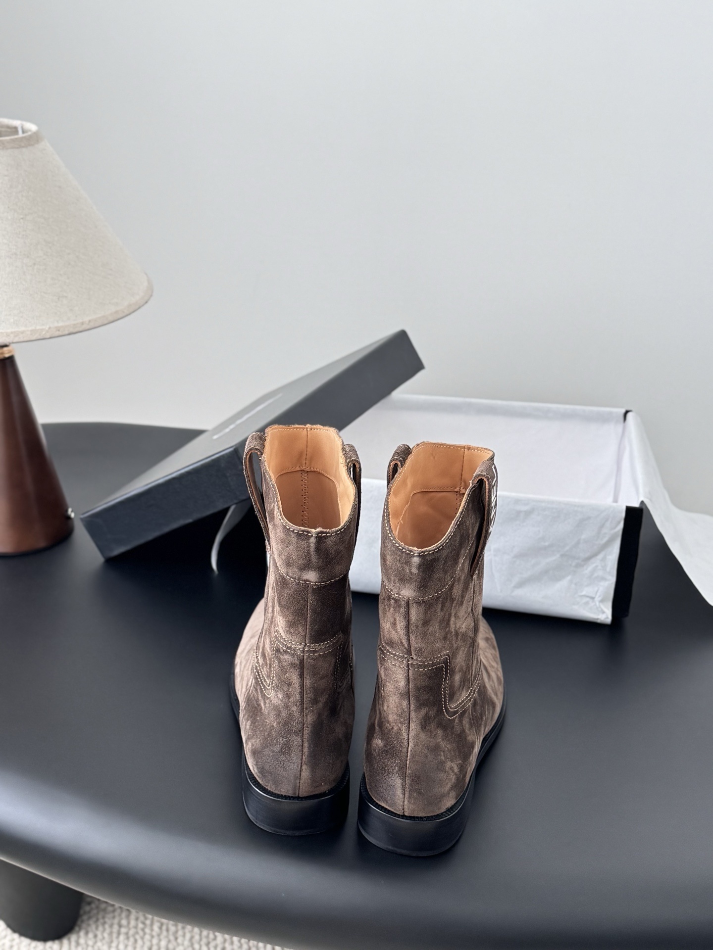 A pair of Chanel brown suede western-style ankle boots with iconic pull tabs and distressed toes on a black table. - Hình 6