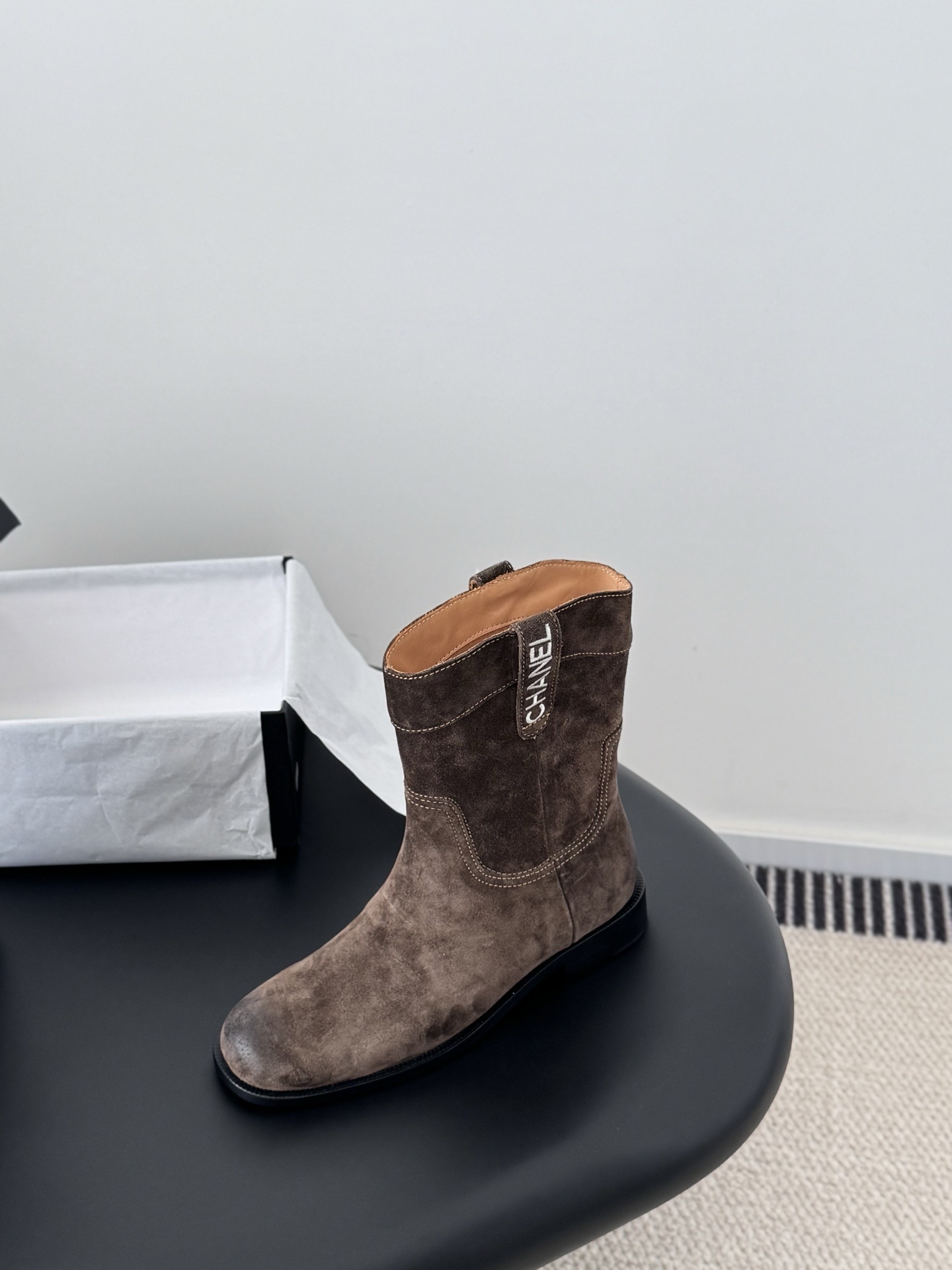 A pair of Chanel brown suede western-style ankle boots with iconic pull tabs and distressed toes on a black table. - Hình 5