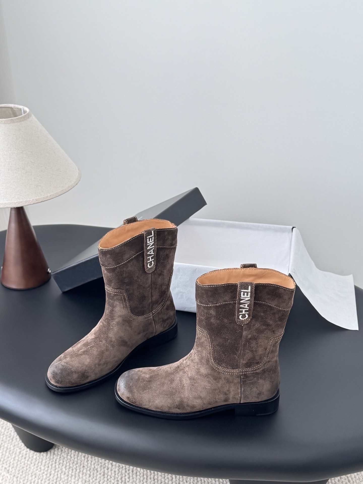 A pair of Chanel brown suede western-style ankle boots with iconic pull tabs and distressed toes on a black table. - Hình 2