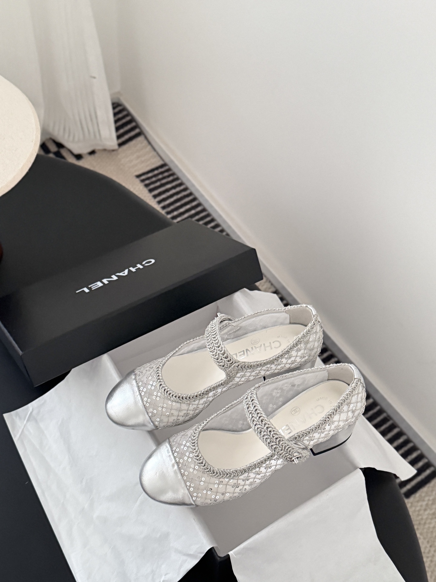 A pair of Chanel silver sequined mesh Mary Jane block heels next to a black Chanel shoe box on a dark surface. - Hình 9