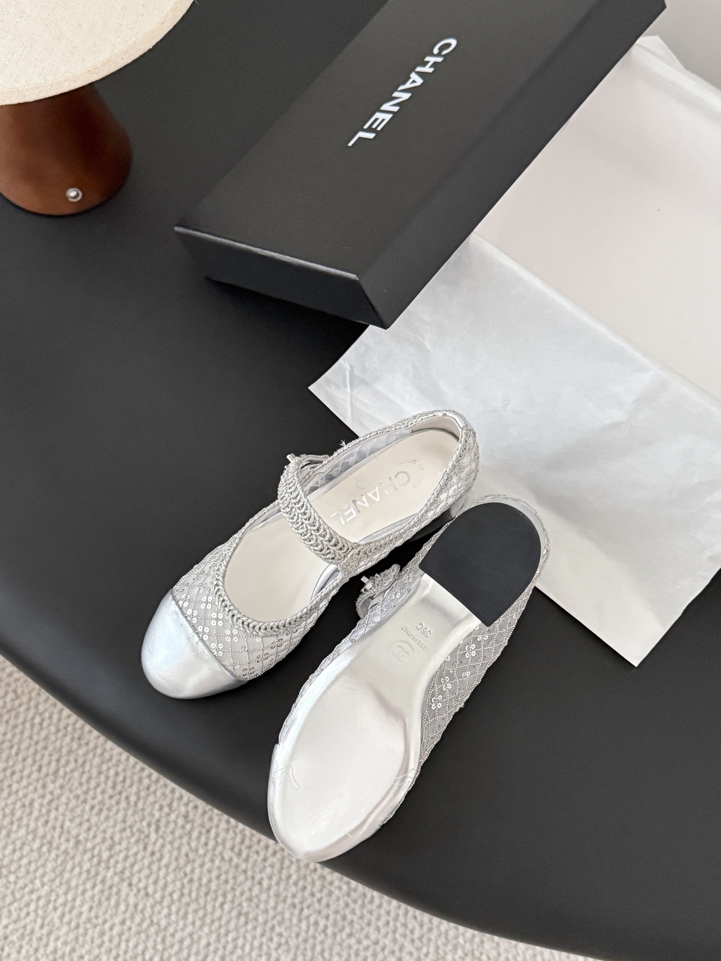 A pair of Chanel silver sequined mesh Mary Jane block heels next to a black Chanel shoe box on a dark surface. - Hình 8