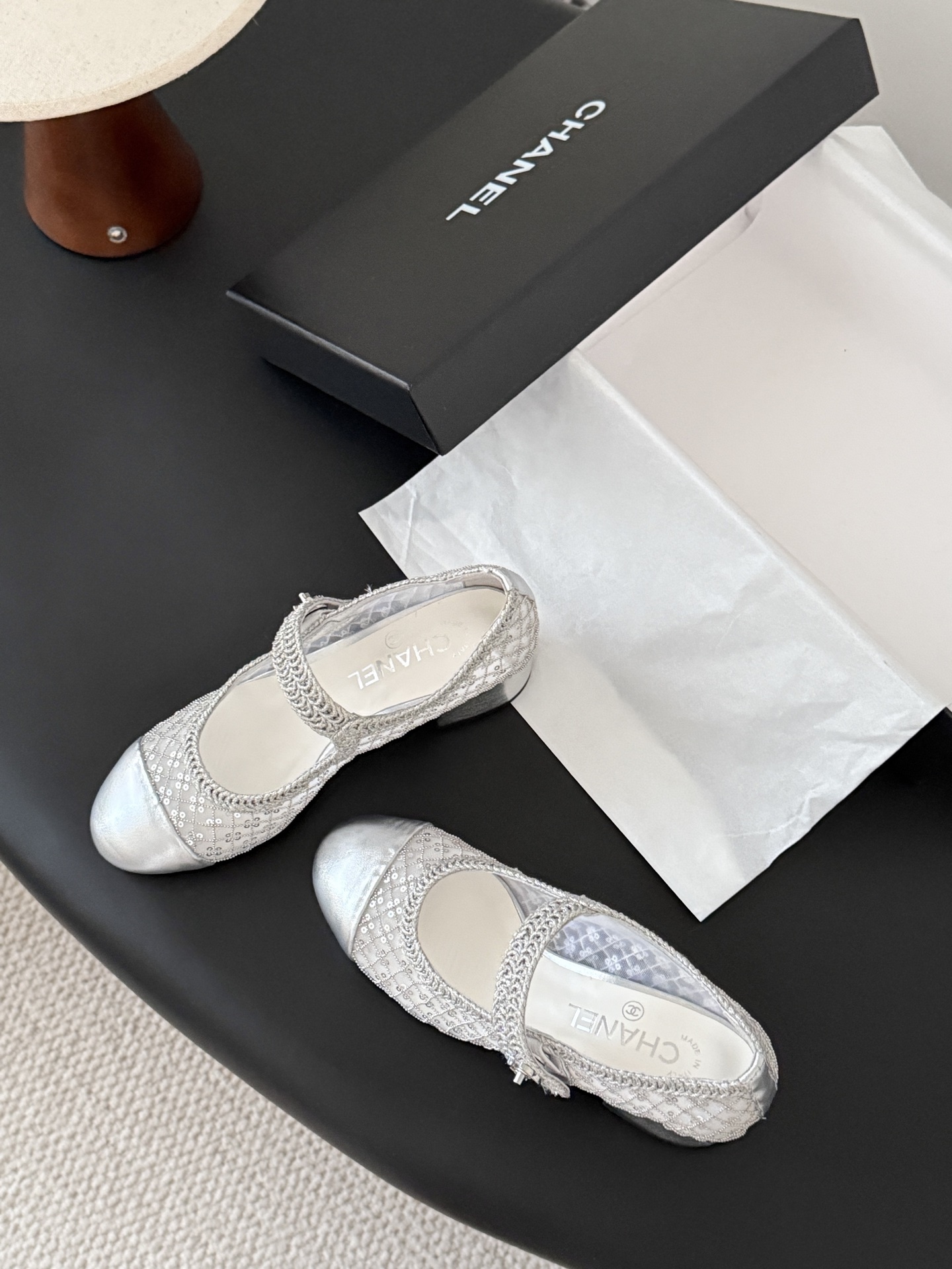 A pair of Chanel silver sequined mesh Mary Jane block heels next to a black Chanel shoe box on a dark surface. - Hình 6