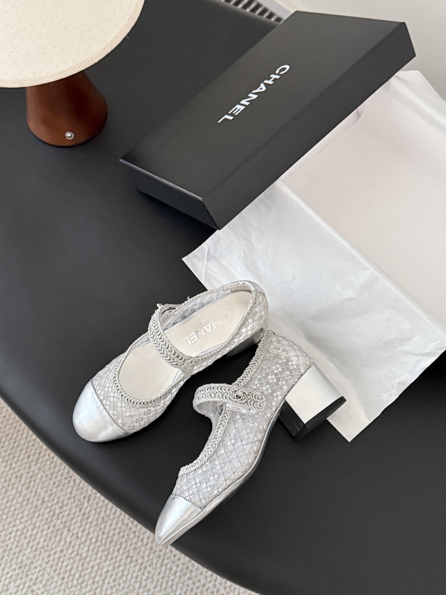 A pair of Chanel silver sequined mesh Mary Jane block heels next to a black Chanel shoe box on a dark surface. - Hình 5