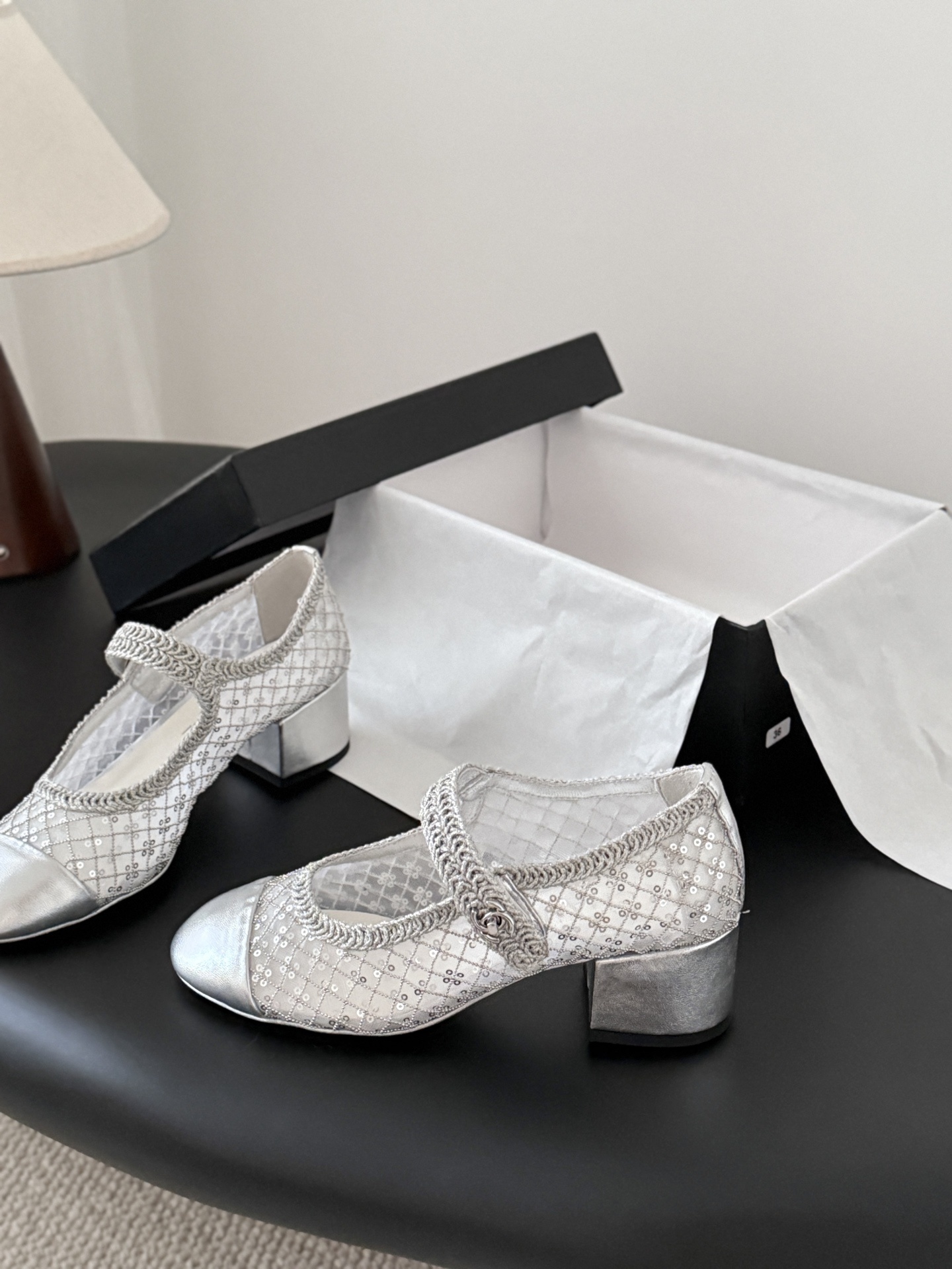 A pair of Chanel silver sequined mesh Mary Jane block heels next to a black Chanel shoe box on a dark surface. - Hình 2