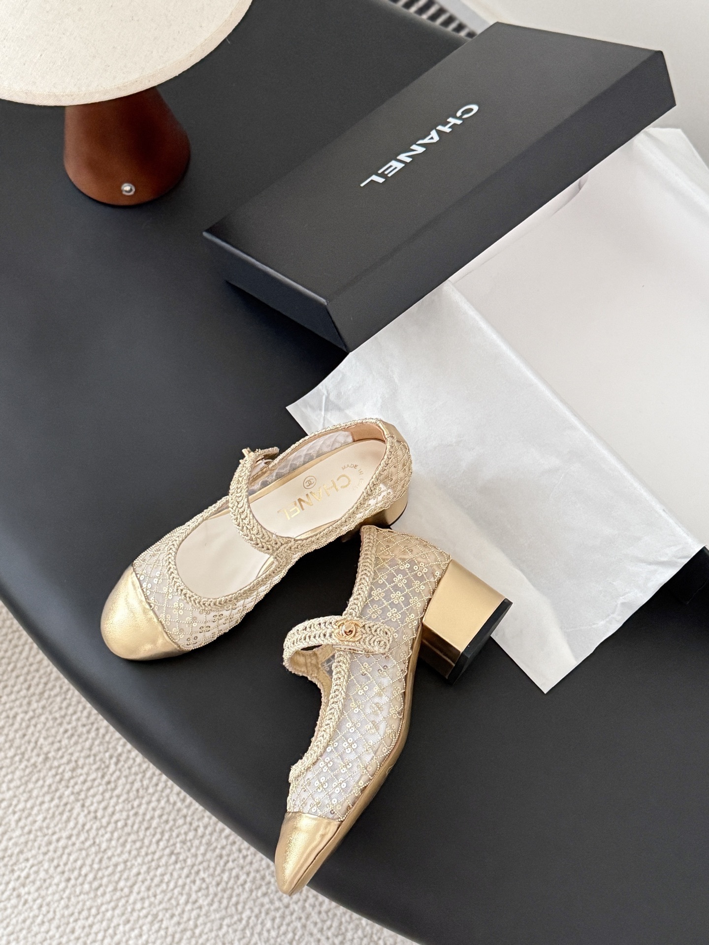 A pair of Chanel gold sequin mesh Mary Jane heels with a gold leather cap toe and block heel on a black table next to a Chanel box. - Hình 8