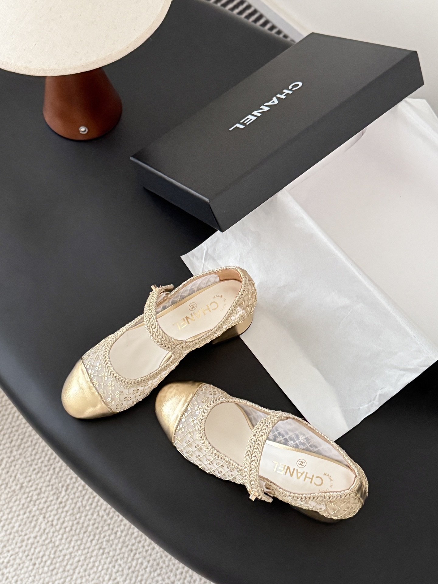 A pair of Chanel gold sequin mesh Mary Jane heels with a gold leather cap toe and block heel on a black table next to a Chanel box. - Hình 4