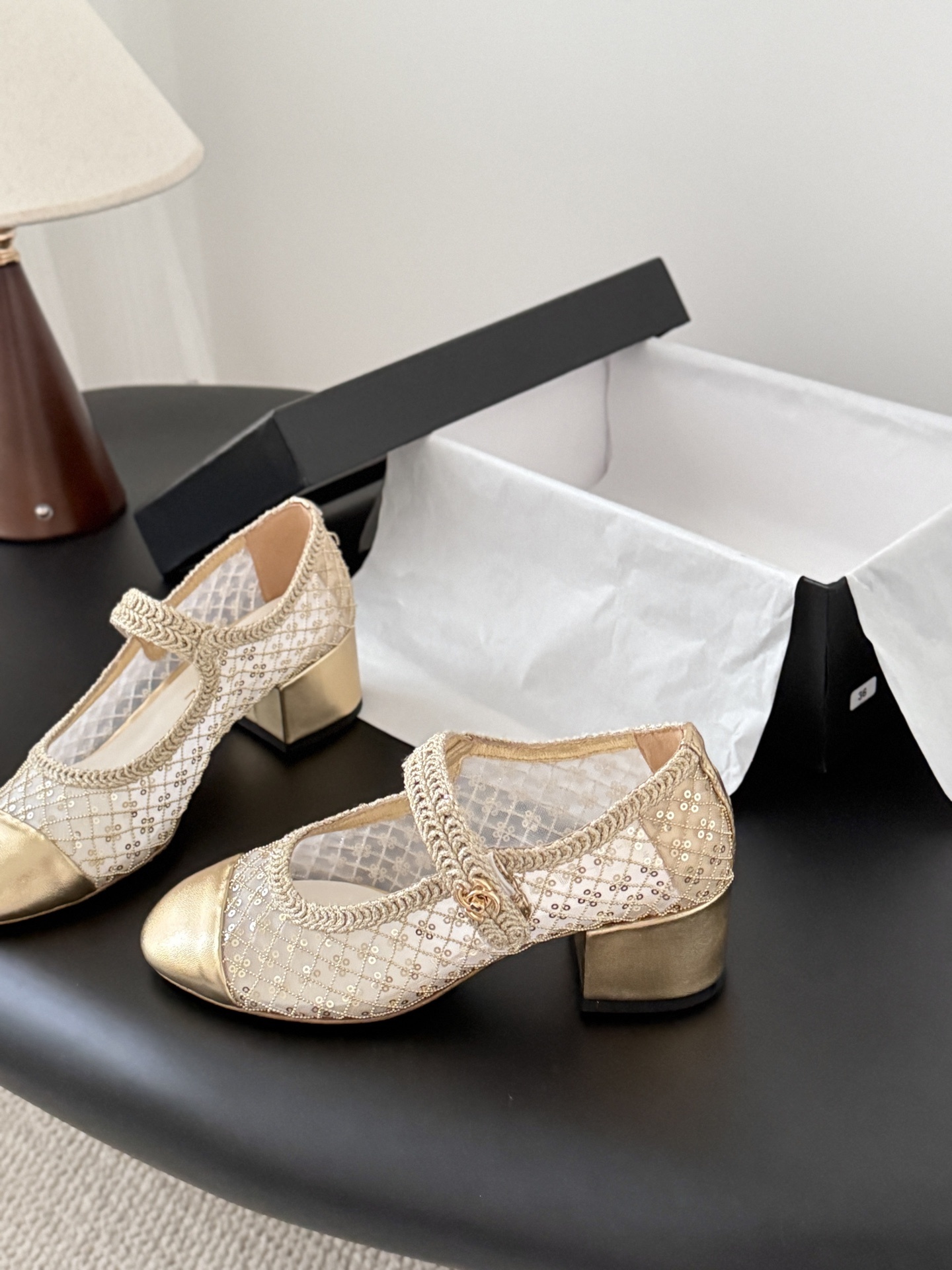 A pair of Chanel gold sequin mesh Mary Jane heels with a gold leather cap toe and block heel on a black table next to a Chanel box. - Hình 2