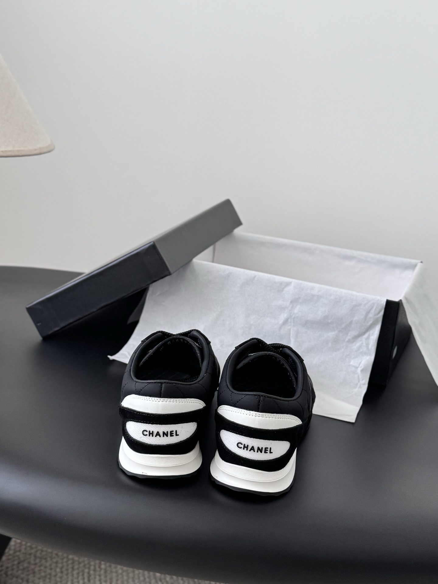 A pair of black Chanel quilted sneakers featuring the CC logo, displayed next to their original black Chanel box. - Hình 6
