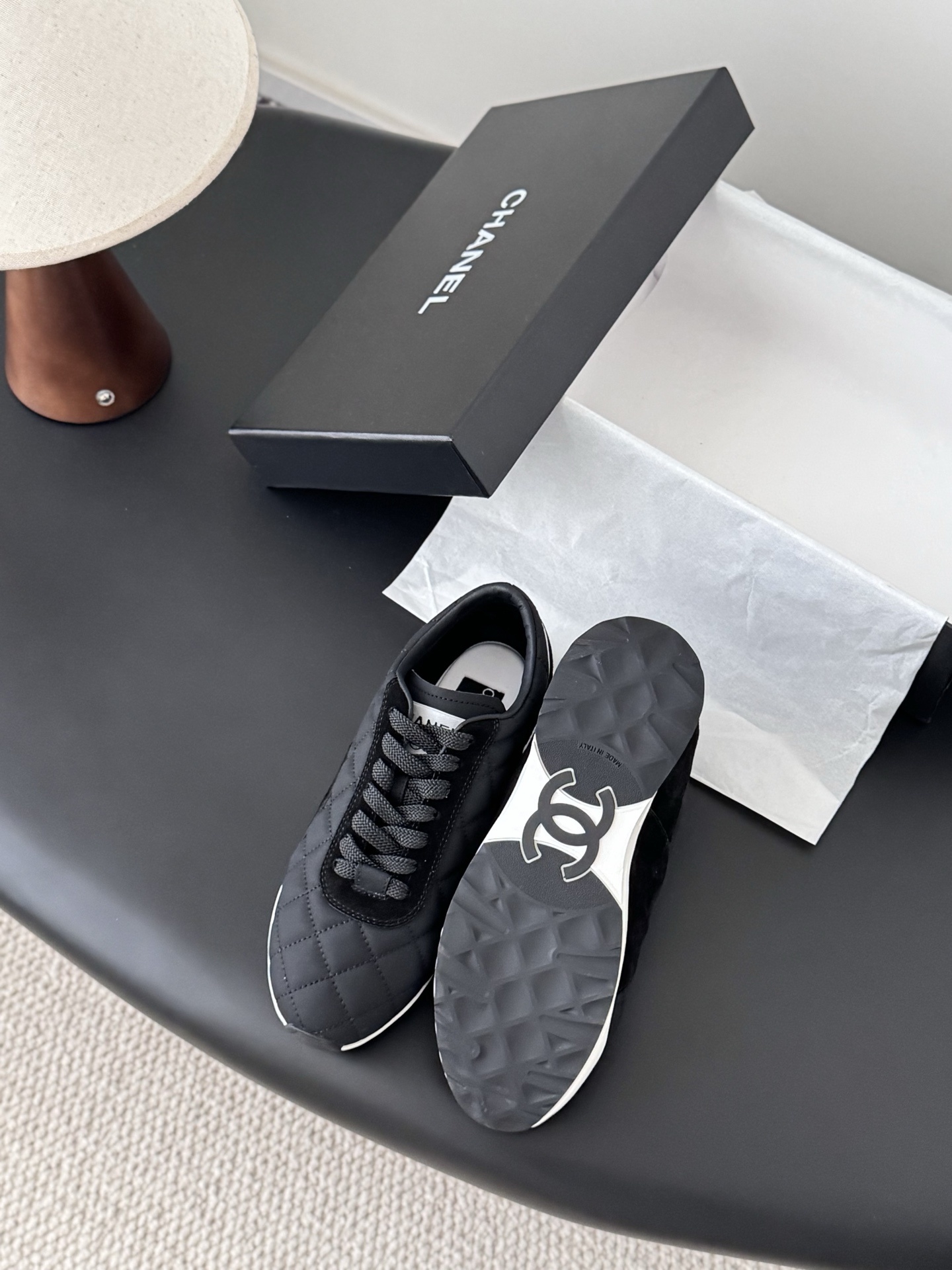 A pair of black Chanel quilted sneakers featuring the CC logo, displayed next to their original black Chanel box. - Hình 4