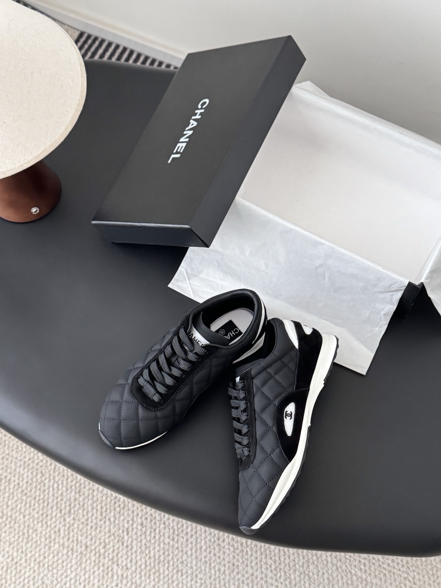 A pair of black Chanel quilted sneakers featuring the CC logo, displayed next to their original black Chanel box. - Hình 3