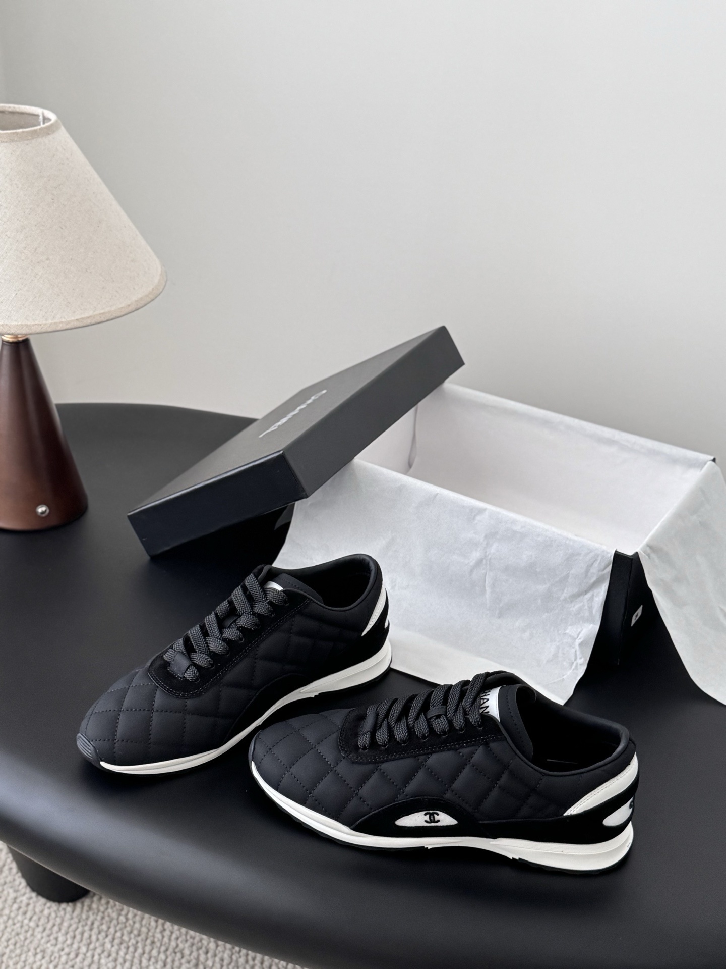 A pair of black Chanel quilted sneakers featuring the CC logo, displayed next to their original black Chanel box. - Hình 2