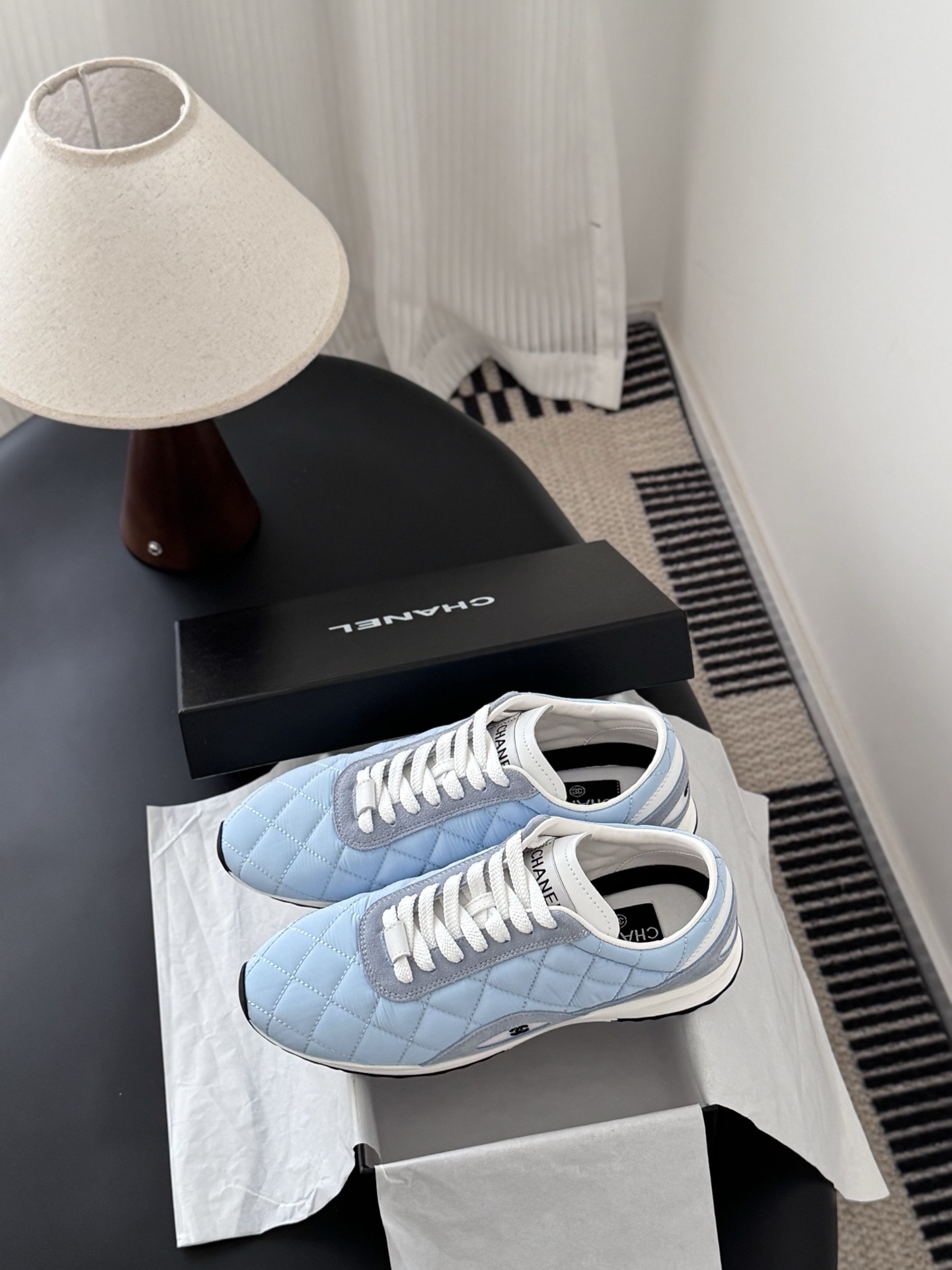 A pair of Chanel light blue quilted sneakers with grey suede accents and white laces presented next to a black Chanel shoe box on a black surface. - Hình 9