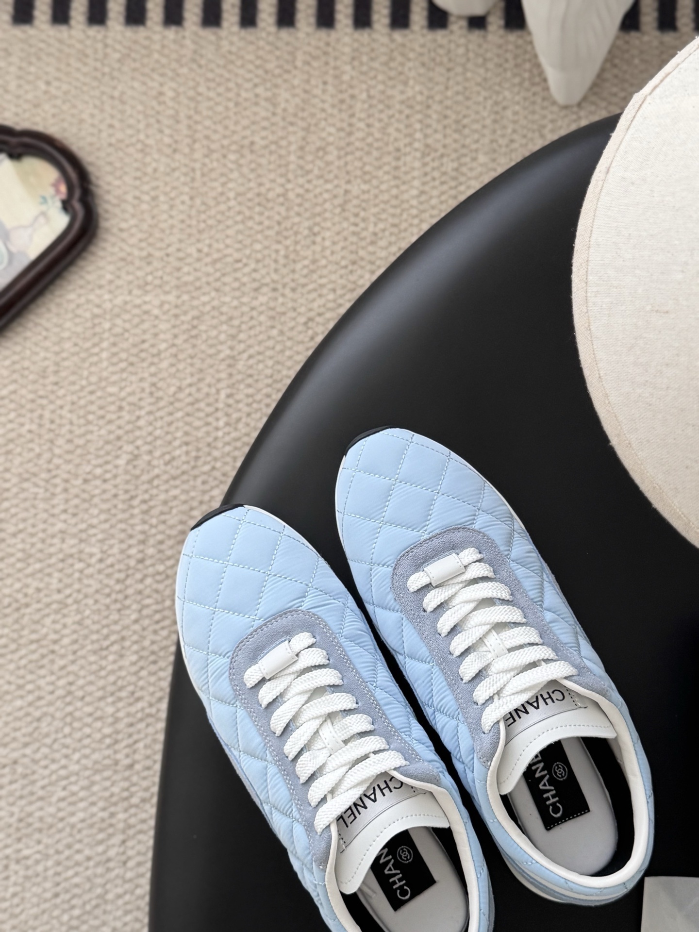 A pair of Chanel light blue quilted sneakers with grey suede accents and white laces presented next to a black Chanel shoe box on a black surface. - Hình 7