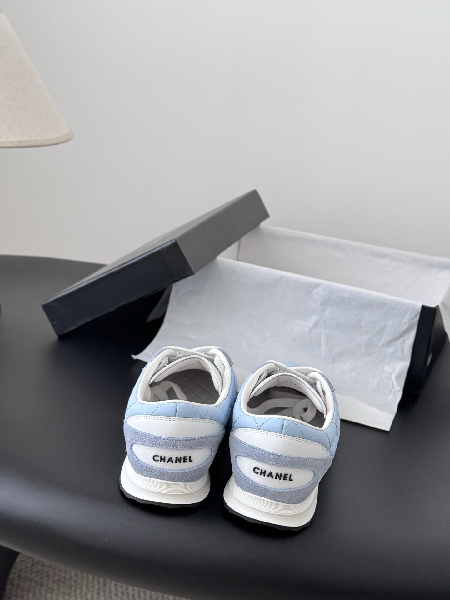 A pair of Chanel light blue quilted sneakers with grey suede accents and white laces presented next to a black Chanel shoe box on a black surface. - Hình 6