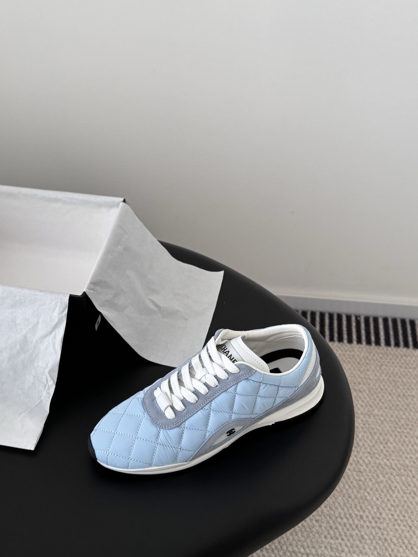 A pair of Chanel light blue quilted sneakers with grey suede accents and white laces presented next to a black Chanel shoe box on a black surface. - Hình 5