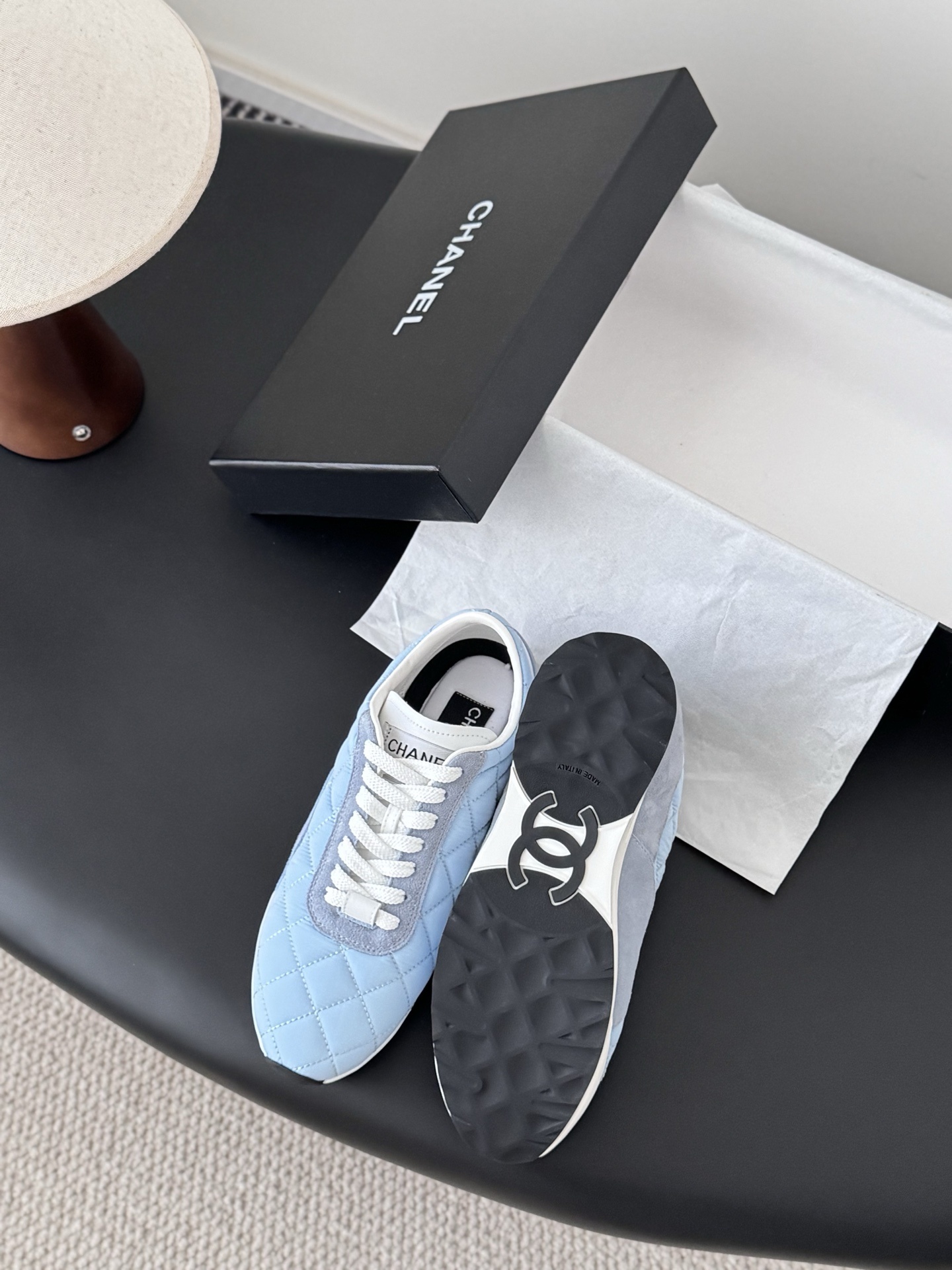 A pair of Chanel light blue quilted sneakers with grey suede accents and white laces presented next to a black Chanel shoe box on a black surface. - Hình 4