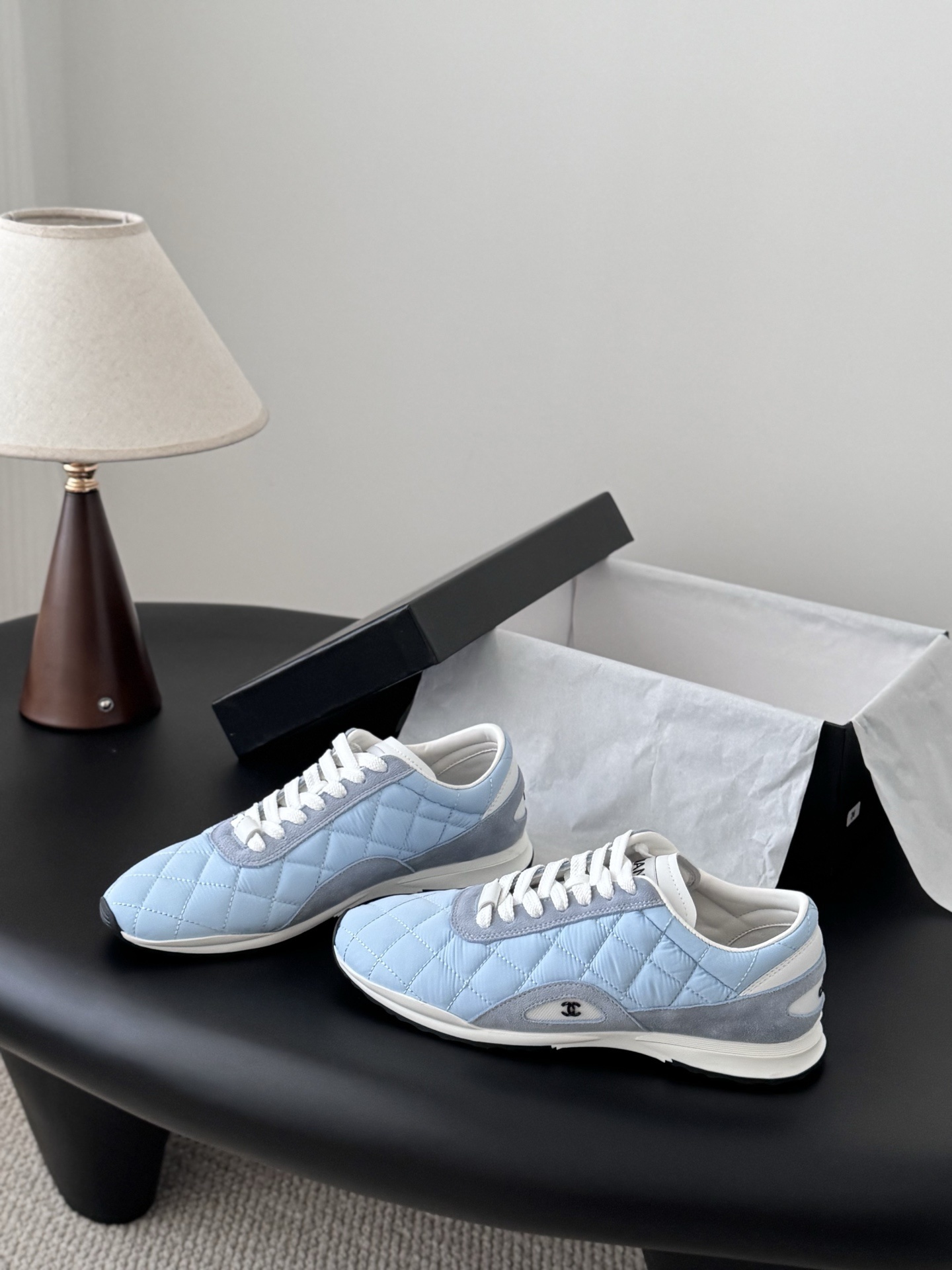 A pair of Chanel light blue quilted sneakers with grey suede accents and white laces presented next to a black Chanel shoe box on a black surface. - Hình 2