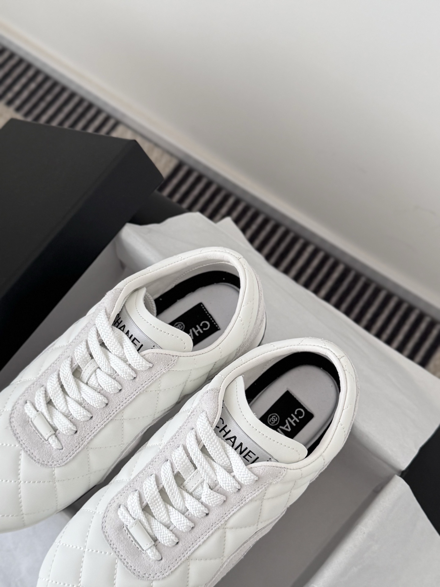 A pair of white Chanel quilted leather sneakers with suede accents next to an original black Chanel shoe box. - Hình 8