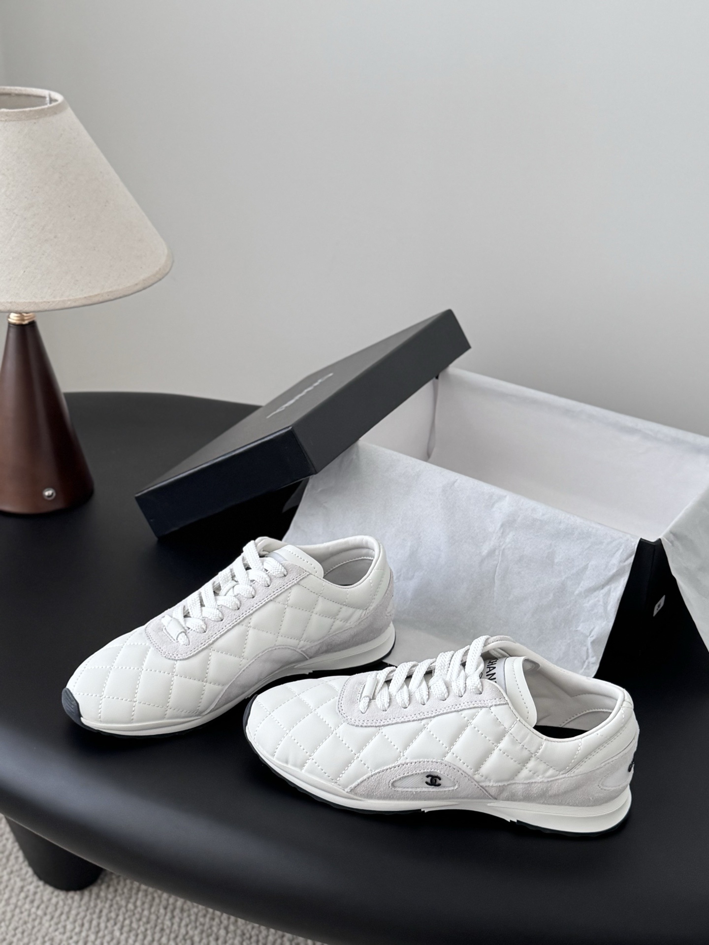 A pair of white Chanel quilted leather sneakers with suede accents next to an original black Chanel shoe box. - Hình 2
