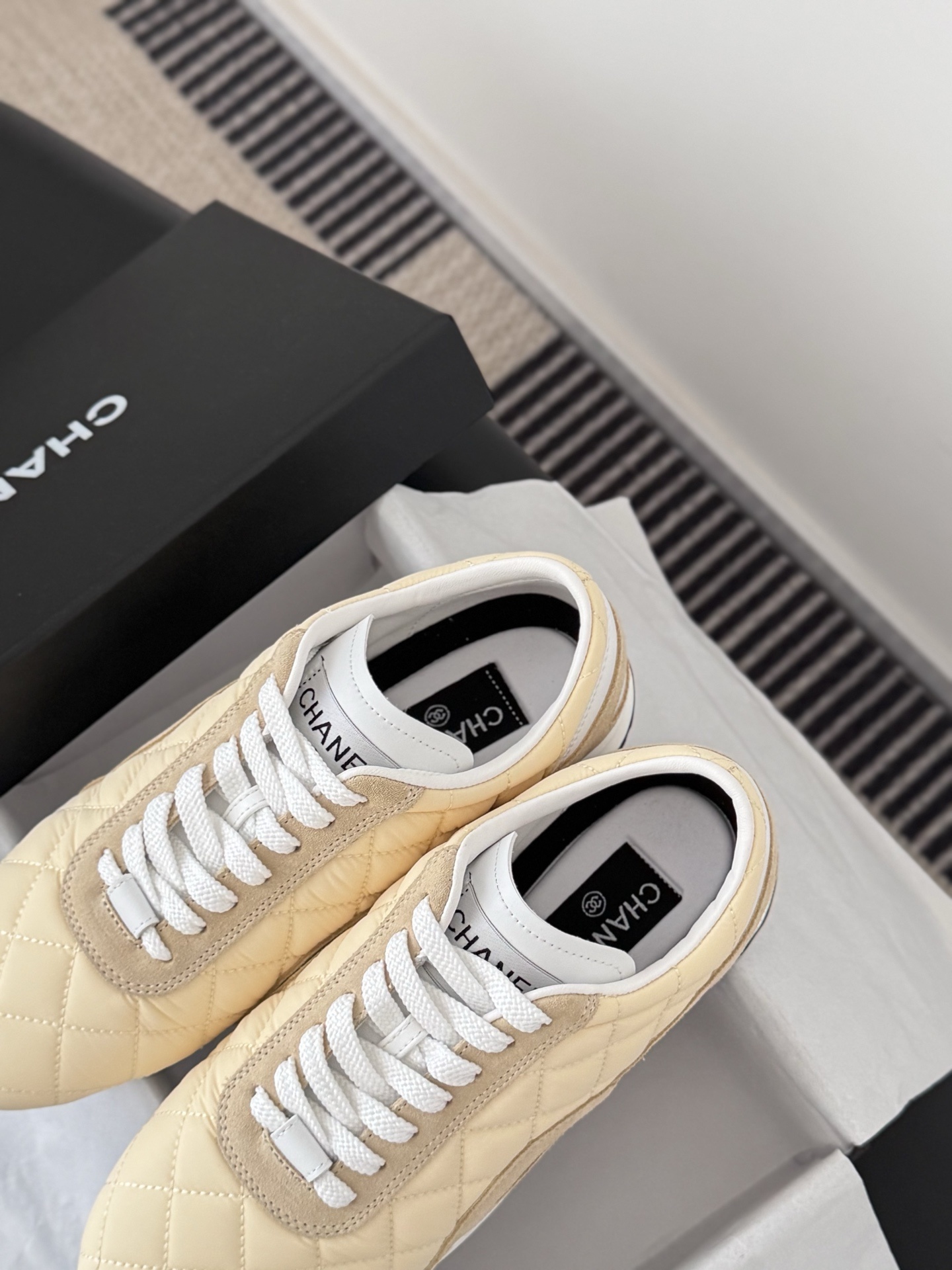 A pair of luxury Chanel sneakers in quilted pale yellow fabric with suede accents and white laces, displayed next to a black Chanel shoebox. - Hình 8