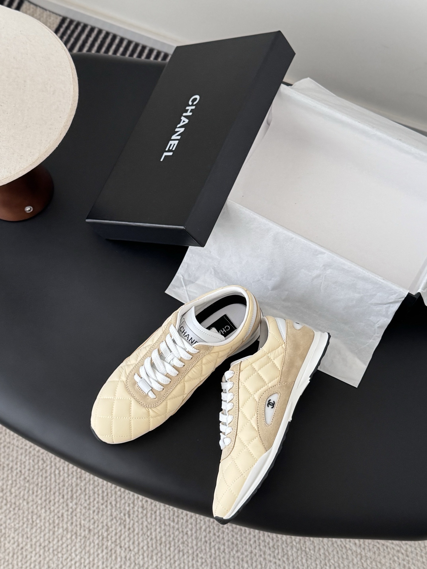 A pair of luxury Chanel sneakers in quilted pale yellow fabric with suede accents and white laces, displayed next to a black Chanel shoebox. - Hình 3