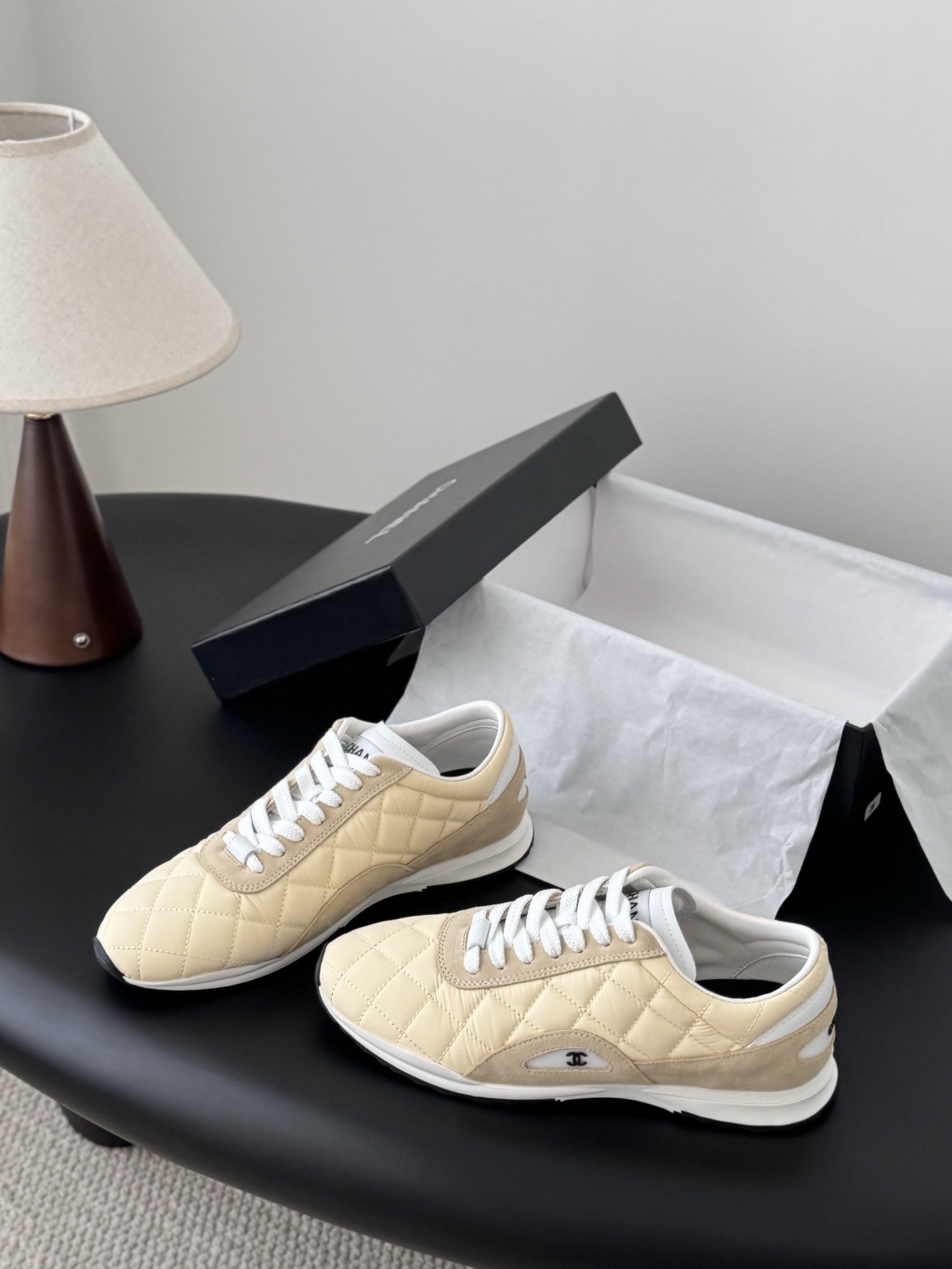 A pair of luxury Chanel sneakers in quilted pale yellow fabric with suede accents and white laces, displayed next to a black Chanel shoebox. - Hình 2