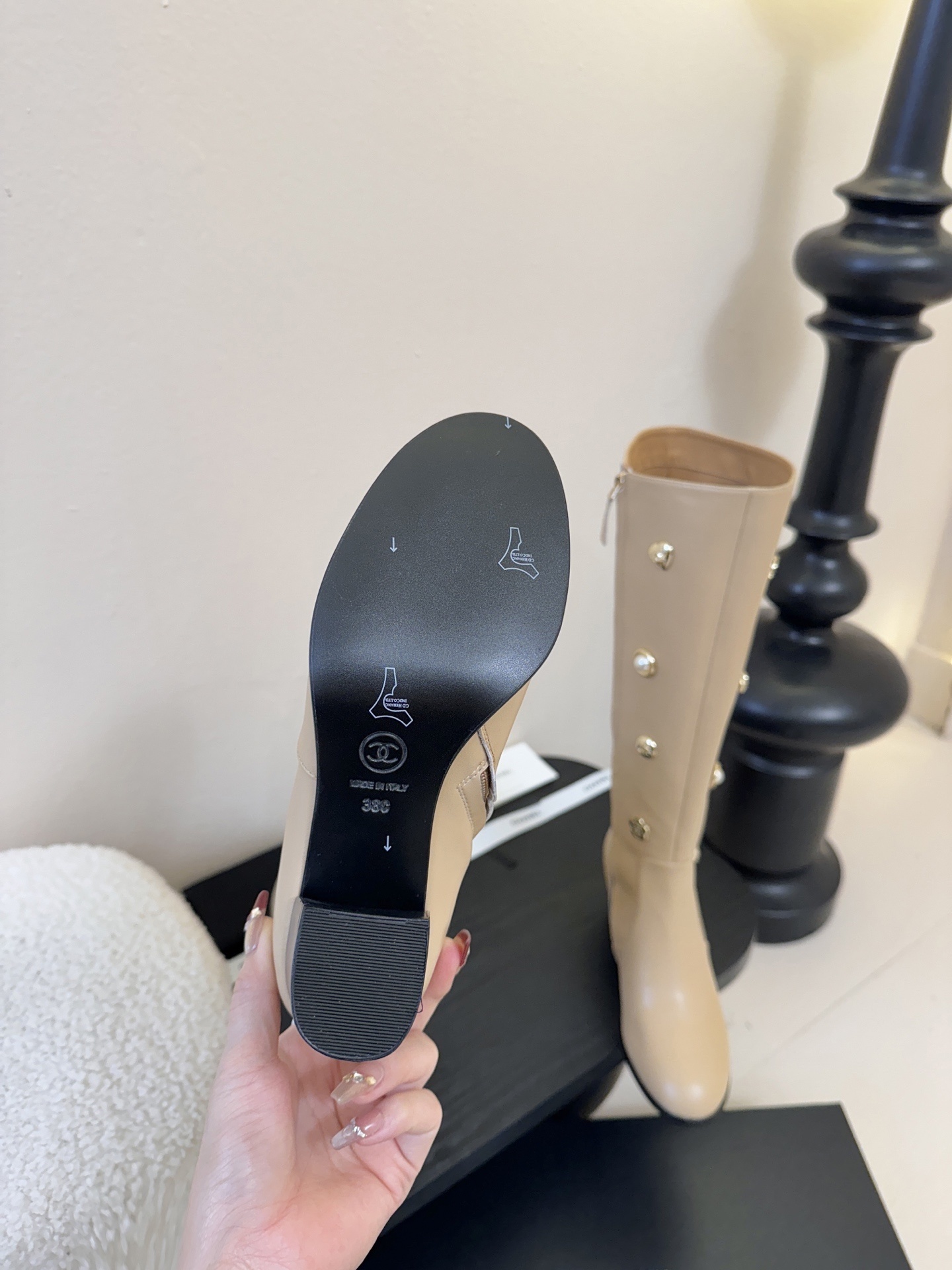 Luxury Chanel beige leather knee-high boots decorated with gold CC logos, pearls, and flower charms on a black display stand. - Hình 9