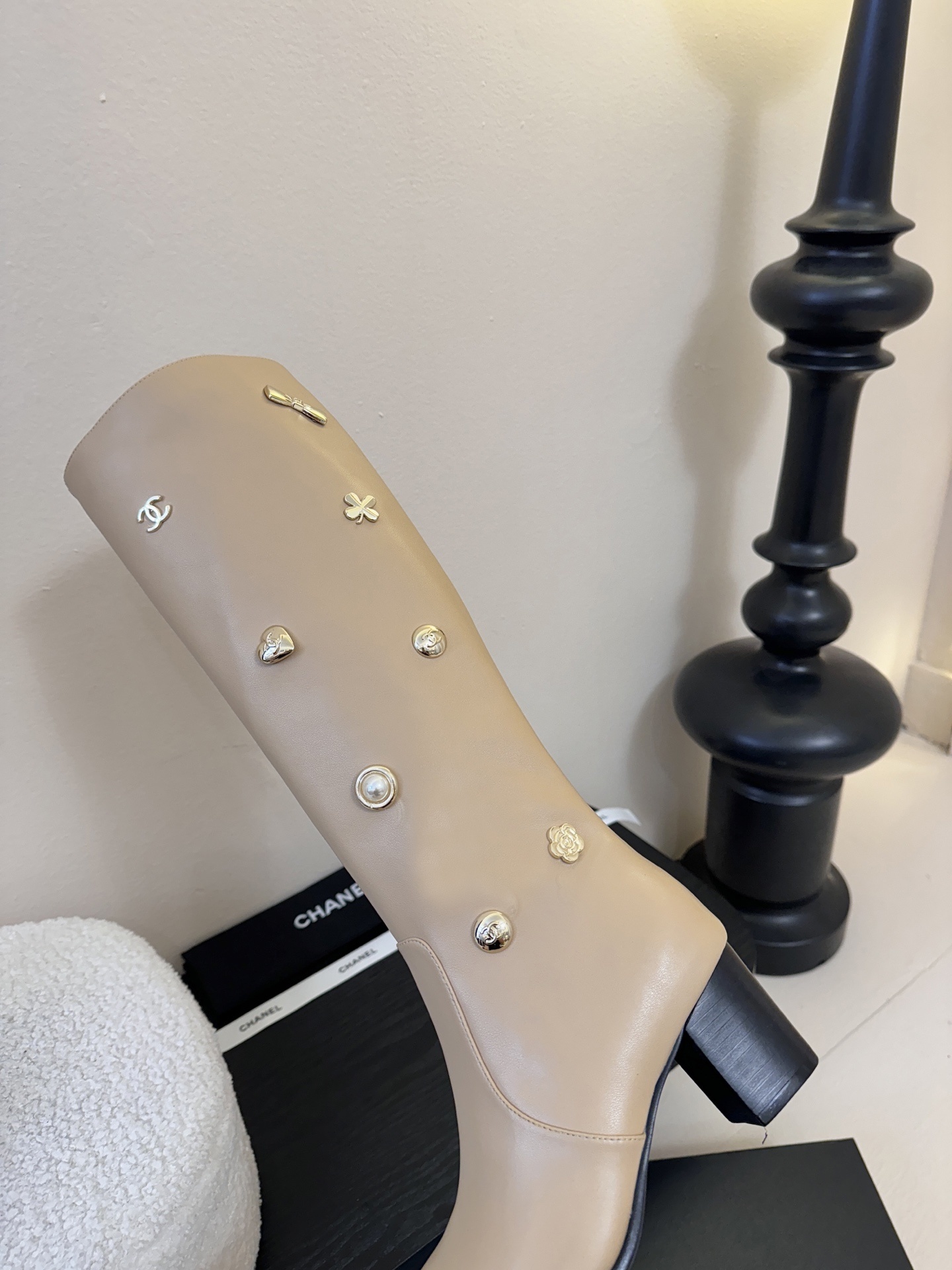 Luxury Chanel beige leather knee-high boots decorated with gold CC logos, pearls, and flower charms on a black display stand. - Hình 8