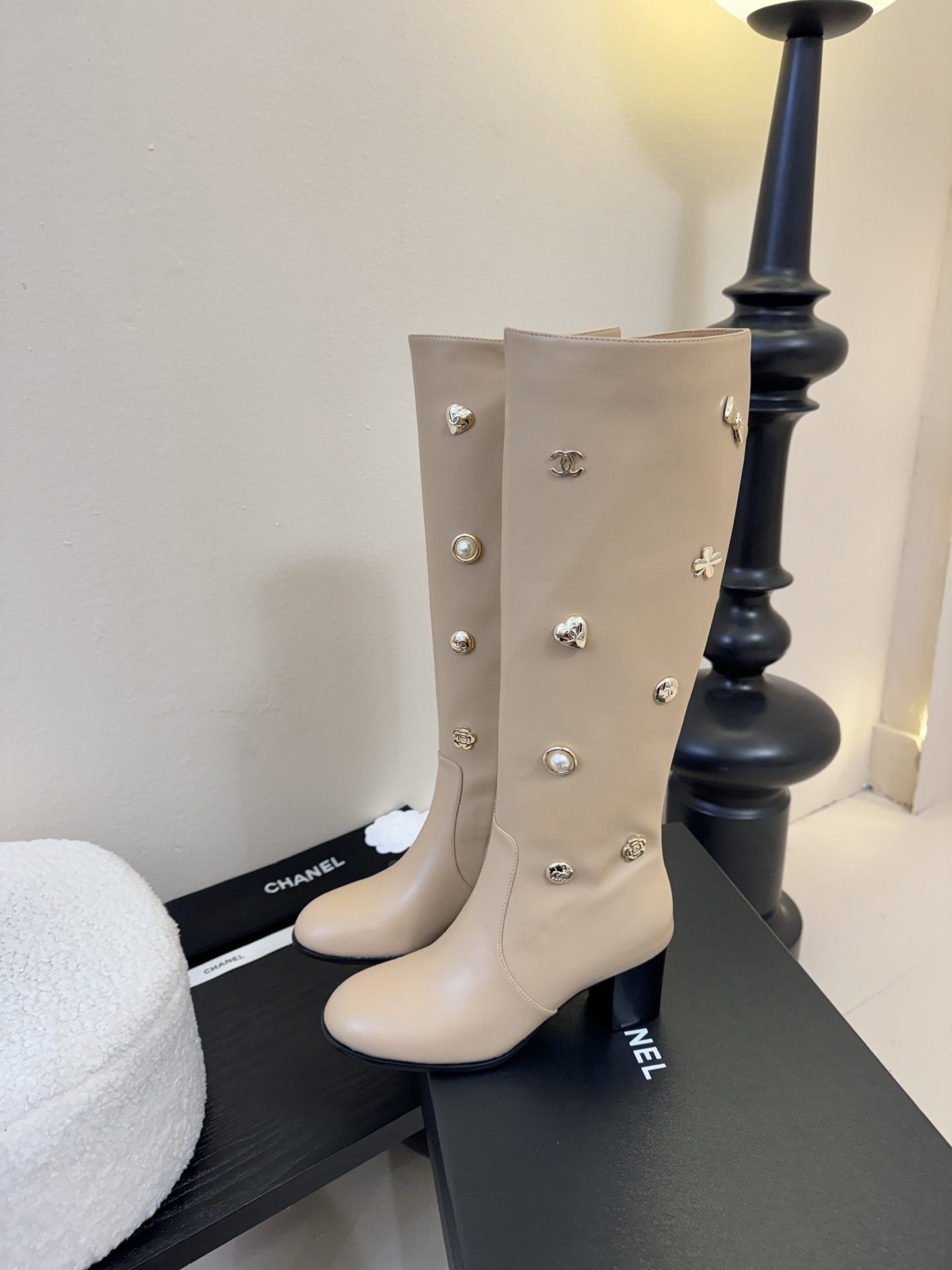 Luxury Chanel beige leather knee-high boots decorated with gold CC logos, pearls, and flower charms on a black display stand. - Hình 5