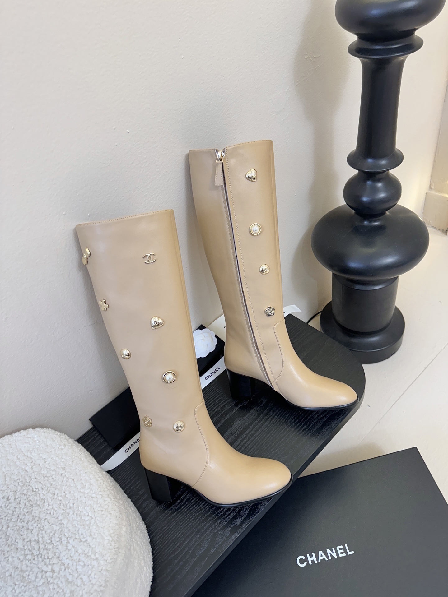Luxury Chanel beige leather knee-high boots decorated with gold CC logos, pearls, and flower charms on a black display stand. - Hình 4