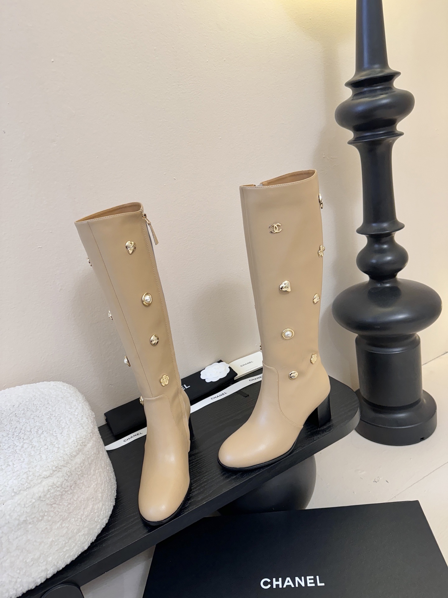 Luxury Chanel beige leather knee-high boots decorated with gold CC logos, pearls, and flower charms on a black display stand. - Hình 3