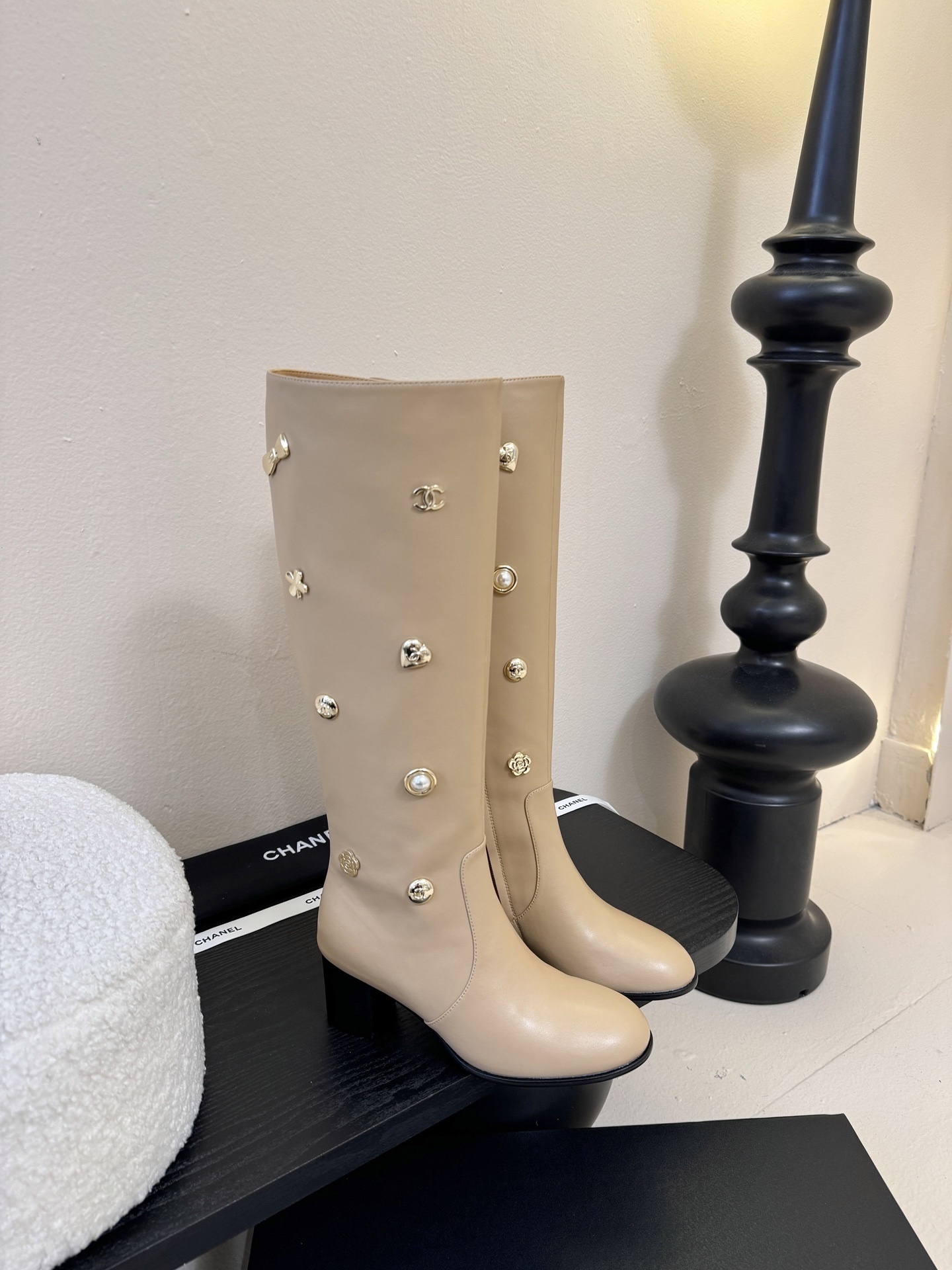 Luxury Chanel beige leather knee-high boots decorated with gold CC logos, pearls, and flower charms on a black display stand. - Hình 2