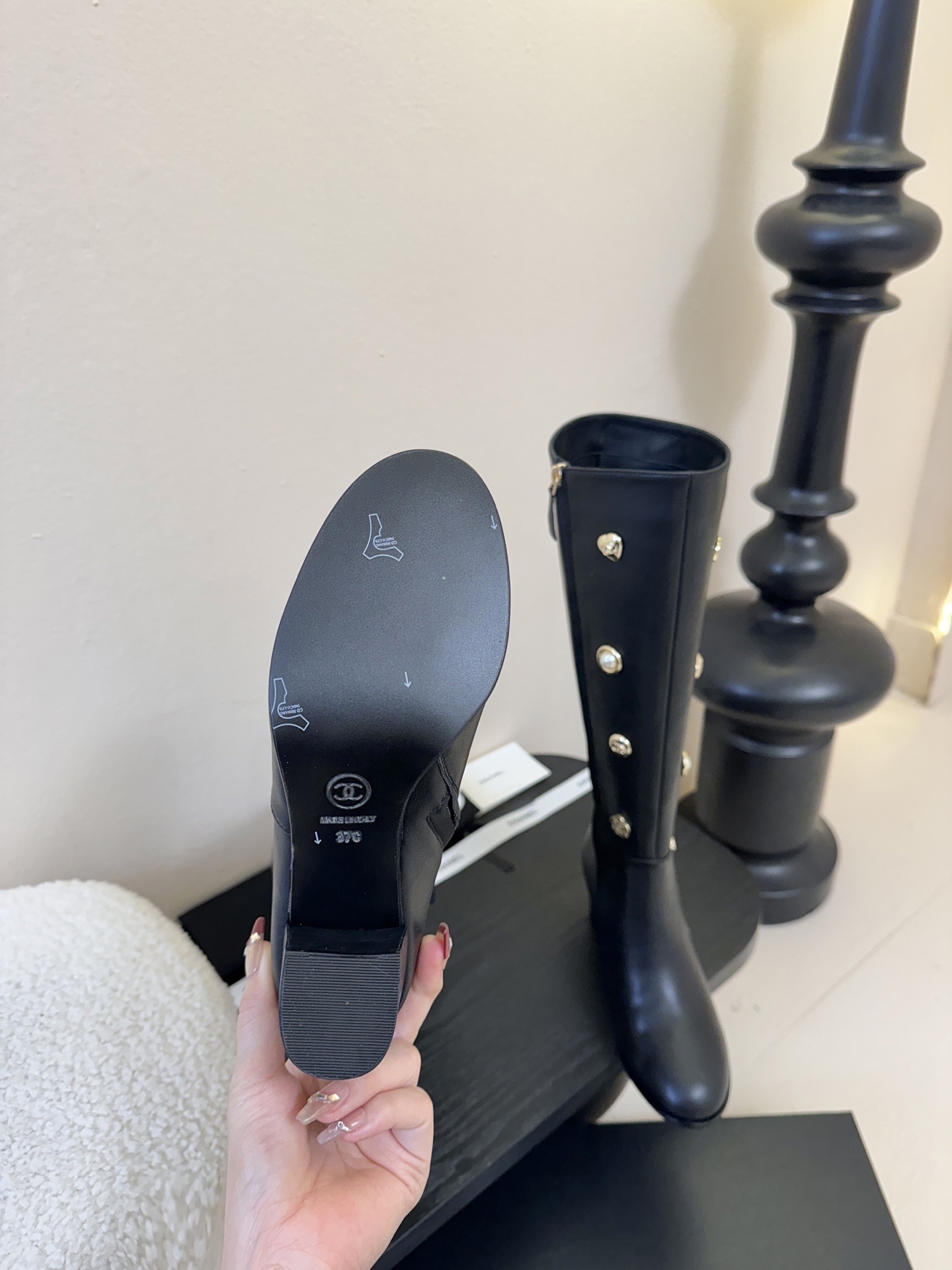 Pair of Chanel black leather knee-high boots decorated with gold CC logos, pearls, and heart charms on a black display. - Hình 9