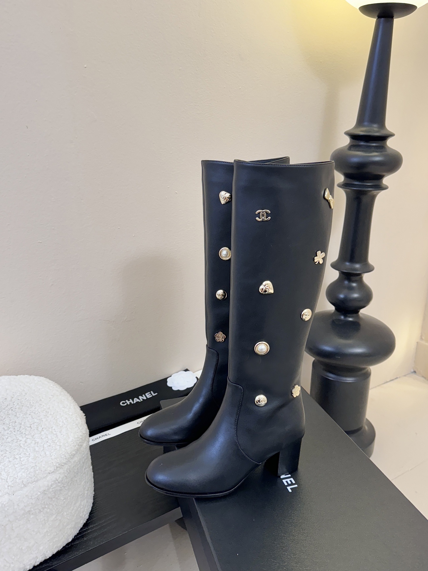 Pair of Chanel black leather knee-high boots decorated with gold CC logos, pearls, and heart charms on a black display. - Hình 5