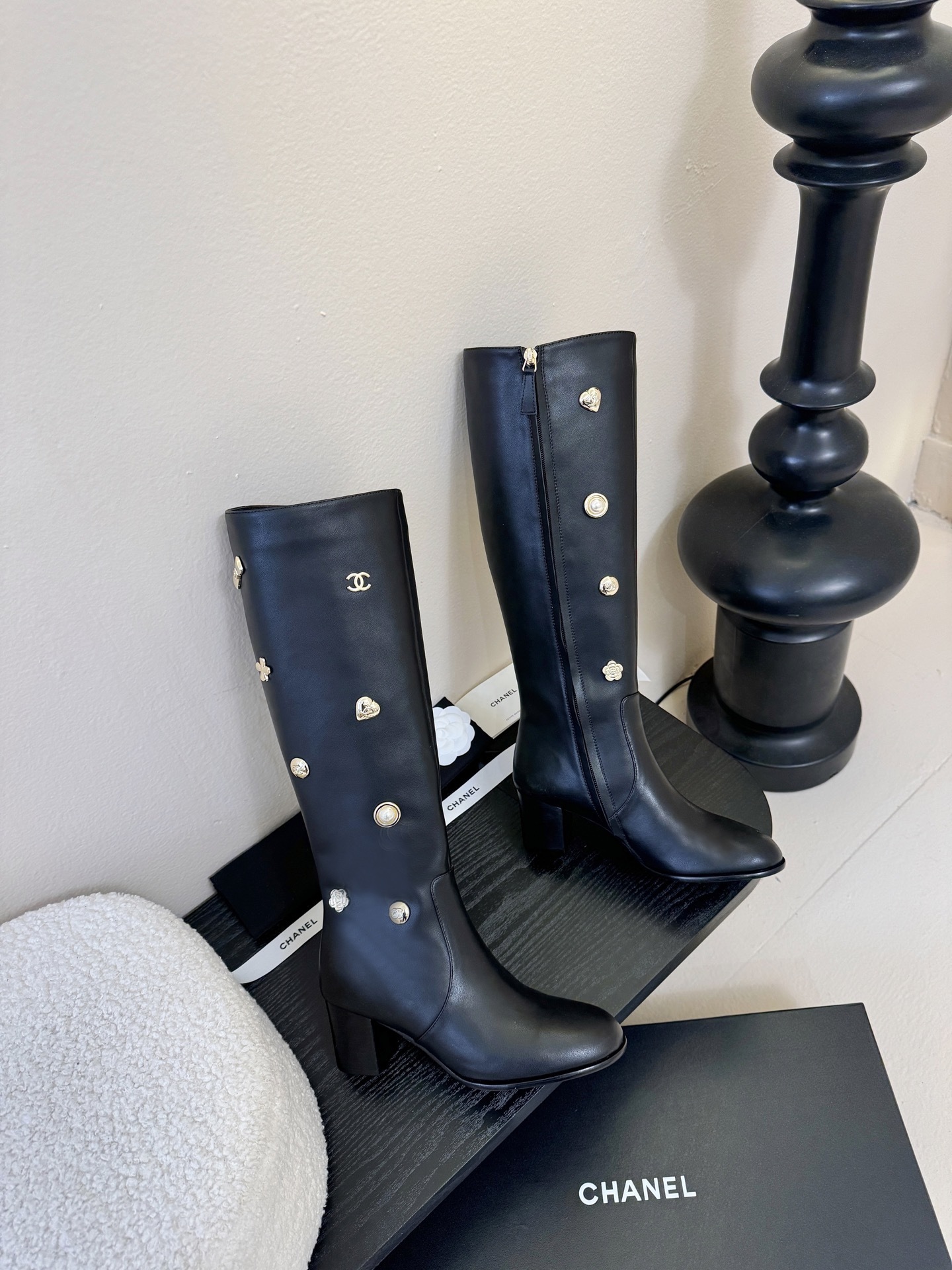 Pair of Chanel black leather knee-high boots decorated with gold CC logos, pearls, and heart charms on a black display. - Hình 4